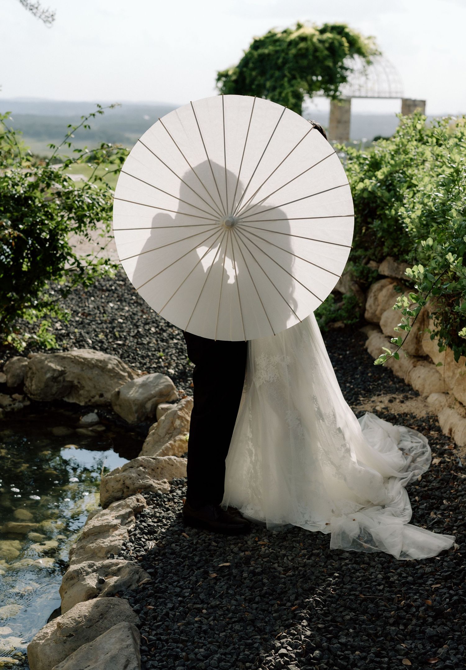 A white paper parasol stands out against black rocks and a white wedding dress beside a small pond in an outdoor garden setting.