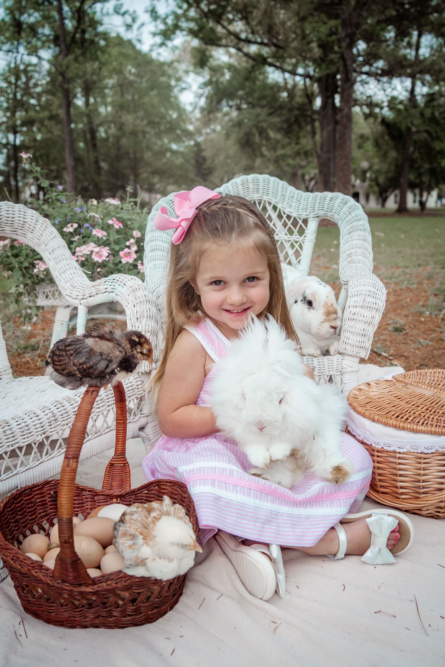A young child in pink dress sits on wicker furniture holding white bunnies with Easter baskets nearby.