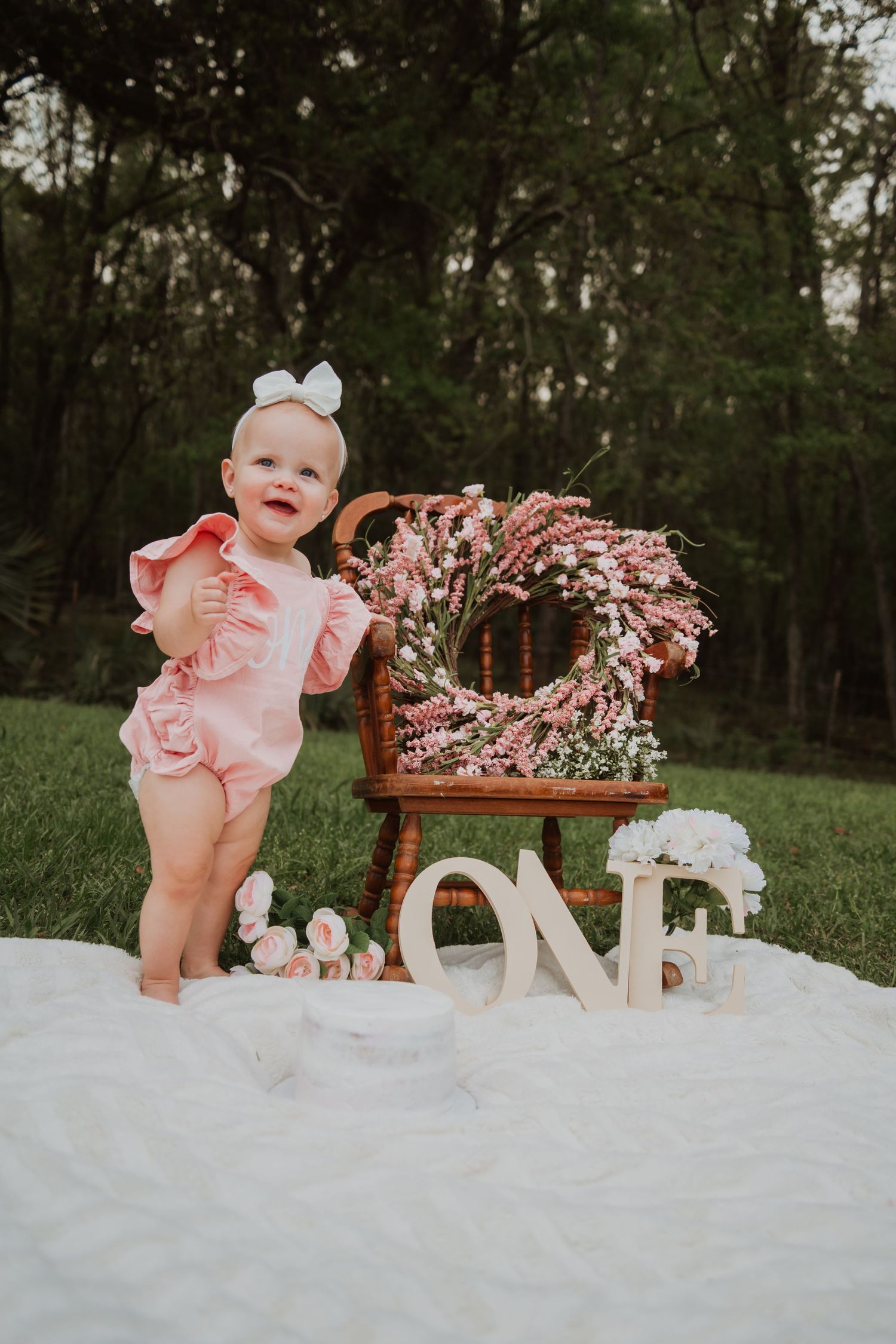 A baby in pink outfit stands beside decorative ONE sign and floral wreath for first birthday photo.