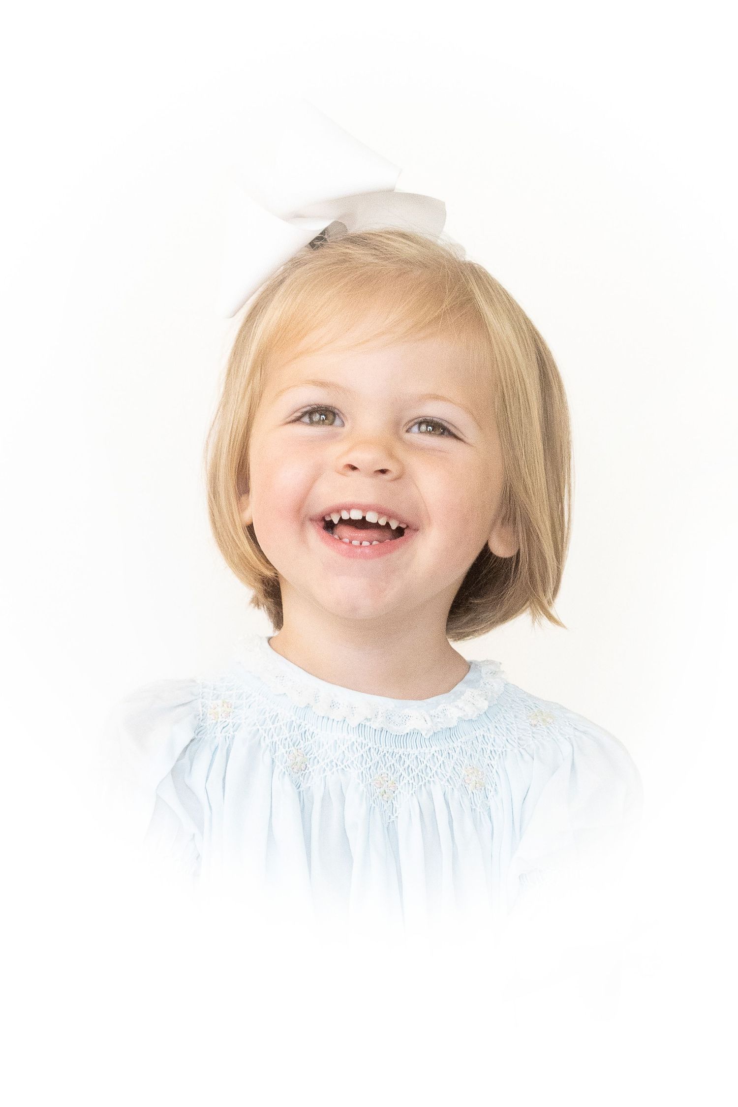 A cheerful toddler wearing a light blue smocked dress smiles brightly against a white background.