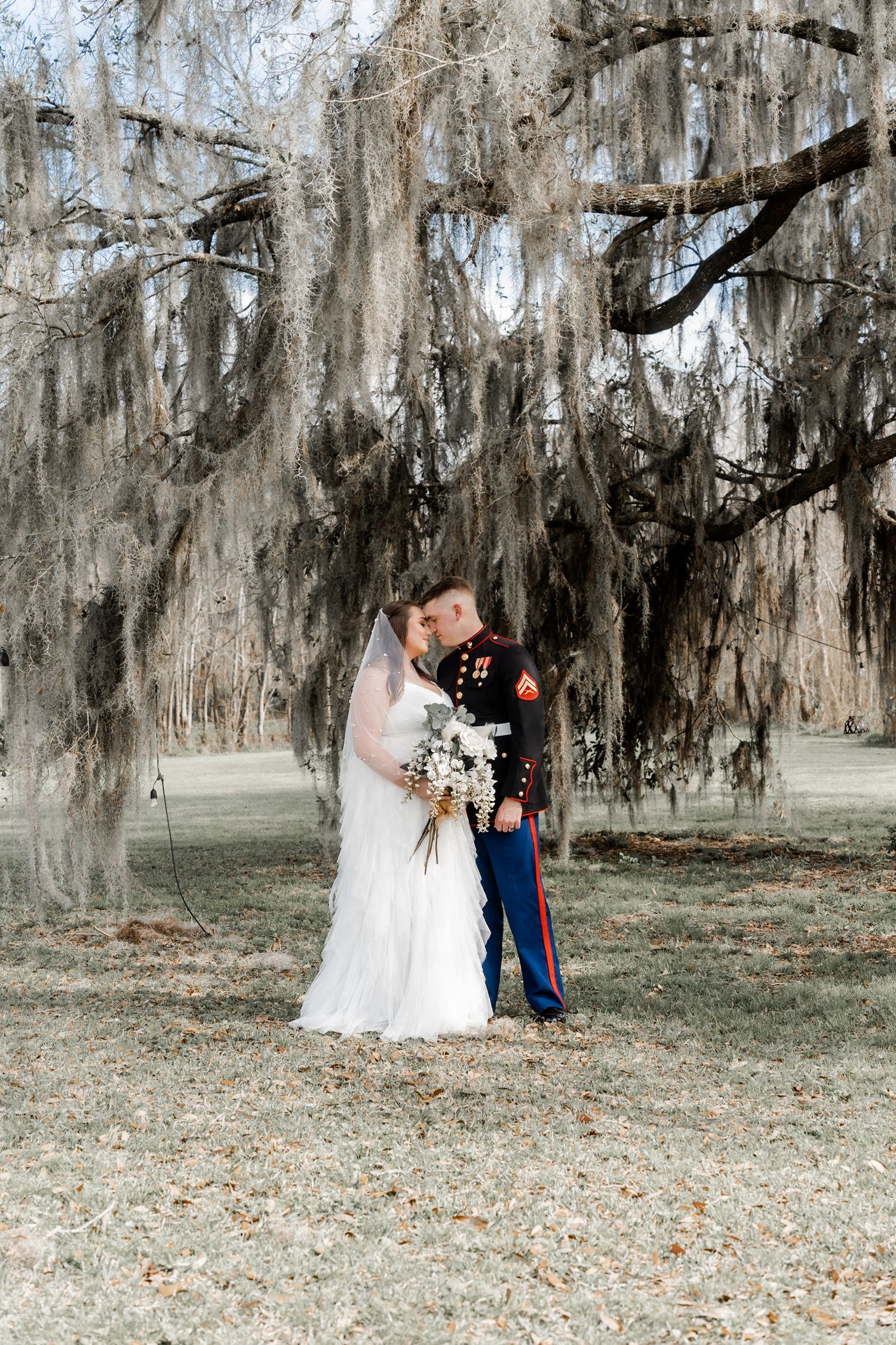 A military wedding couple poses under dramatic hanging Spanish moss in a scenic outdoor setting.