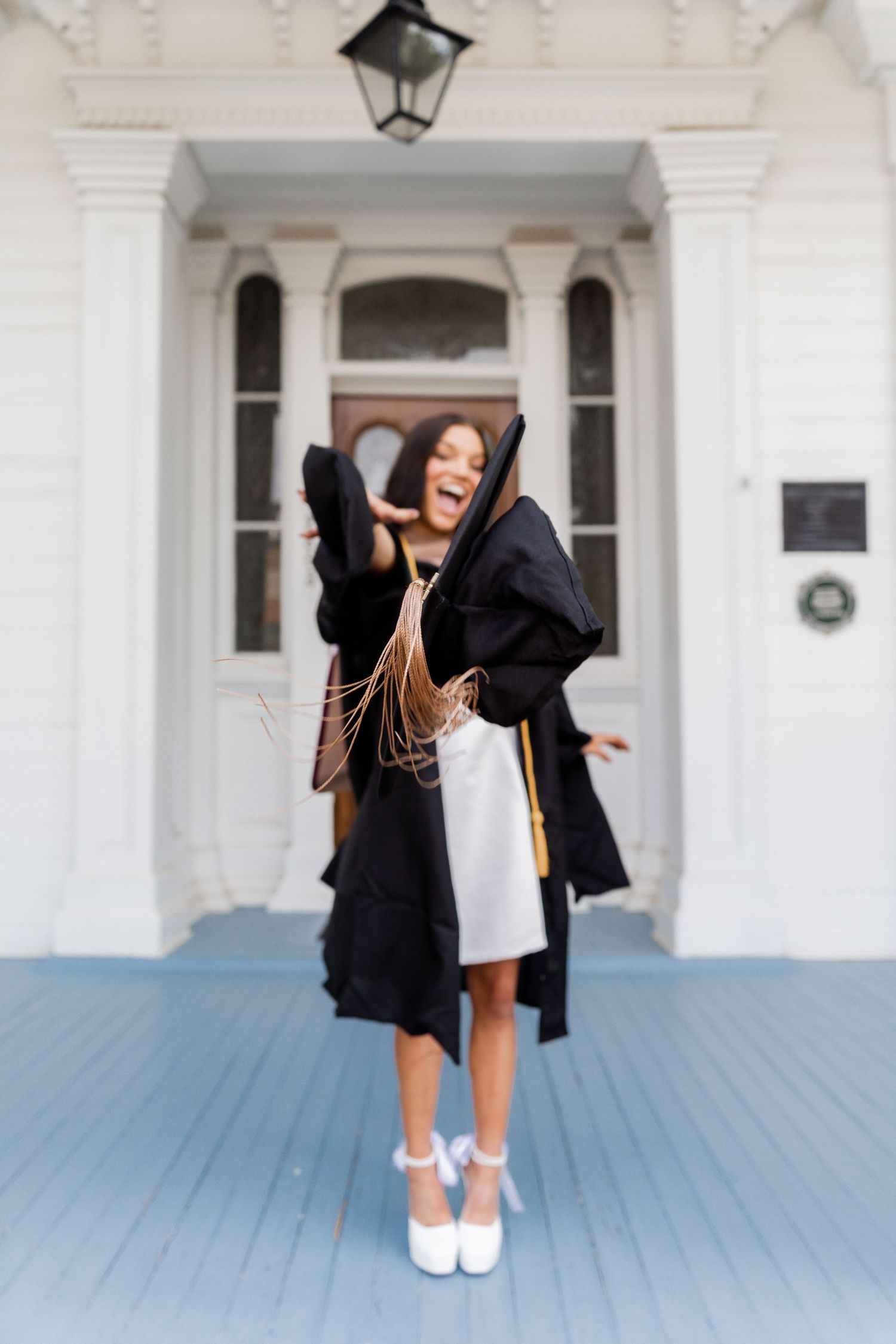 Graduate in black robe and white dress jumps joyfully on blue porch in celebration.