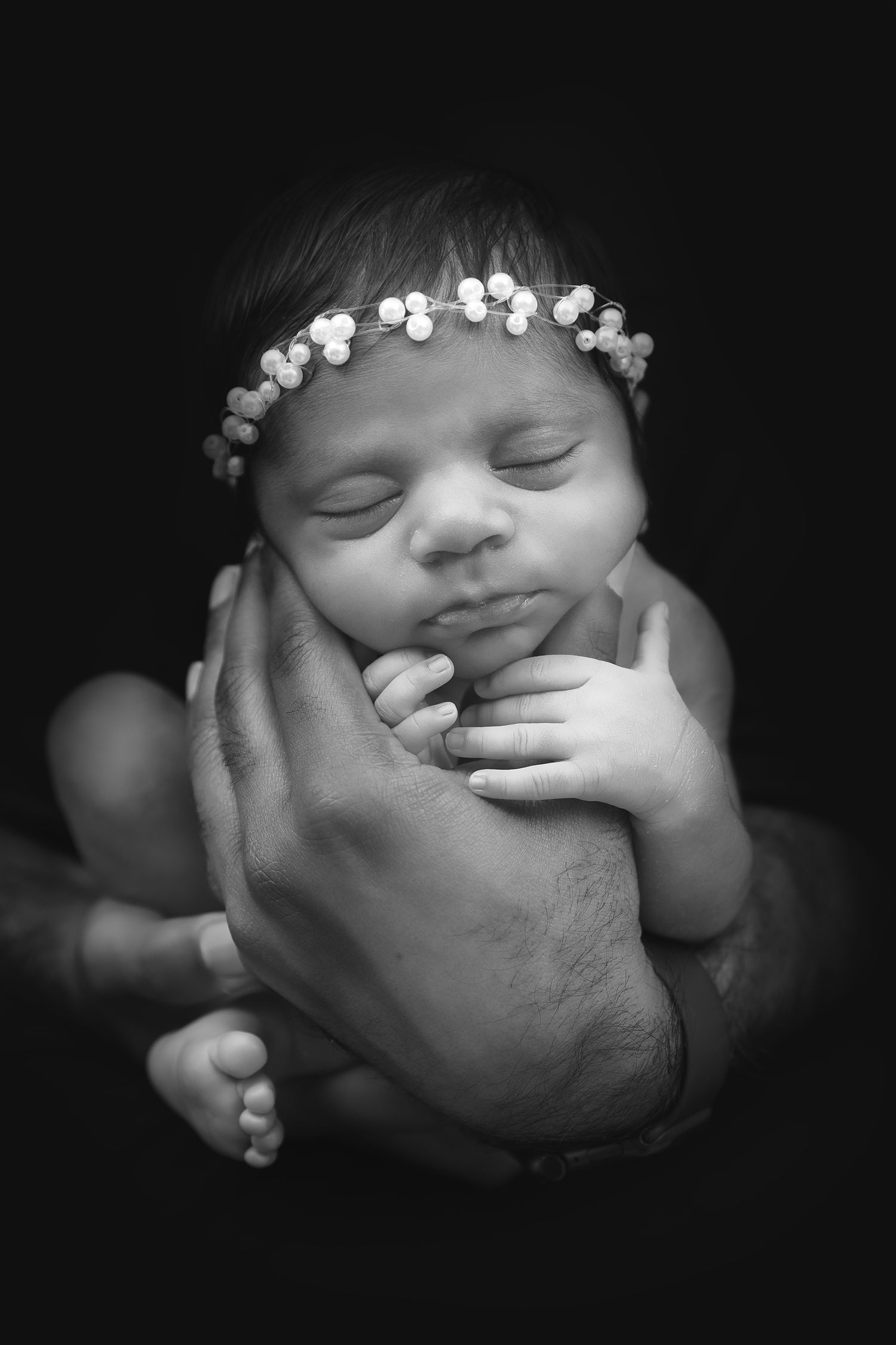 Black and white newborn baby girl sleeping in parent's hands wearing a delicate floral crown.