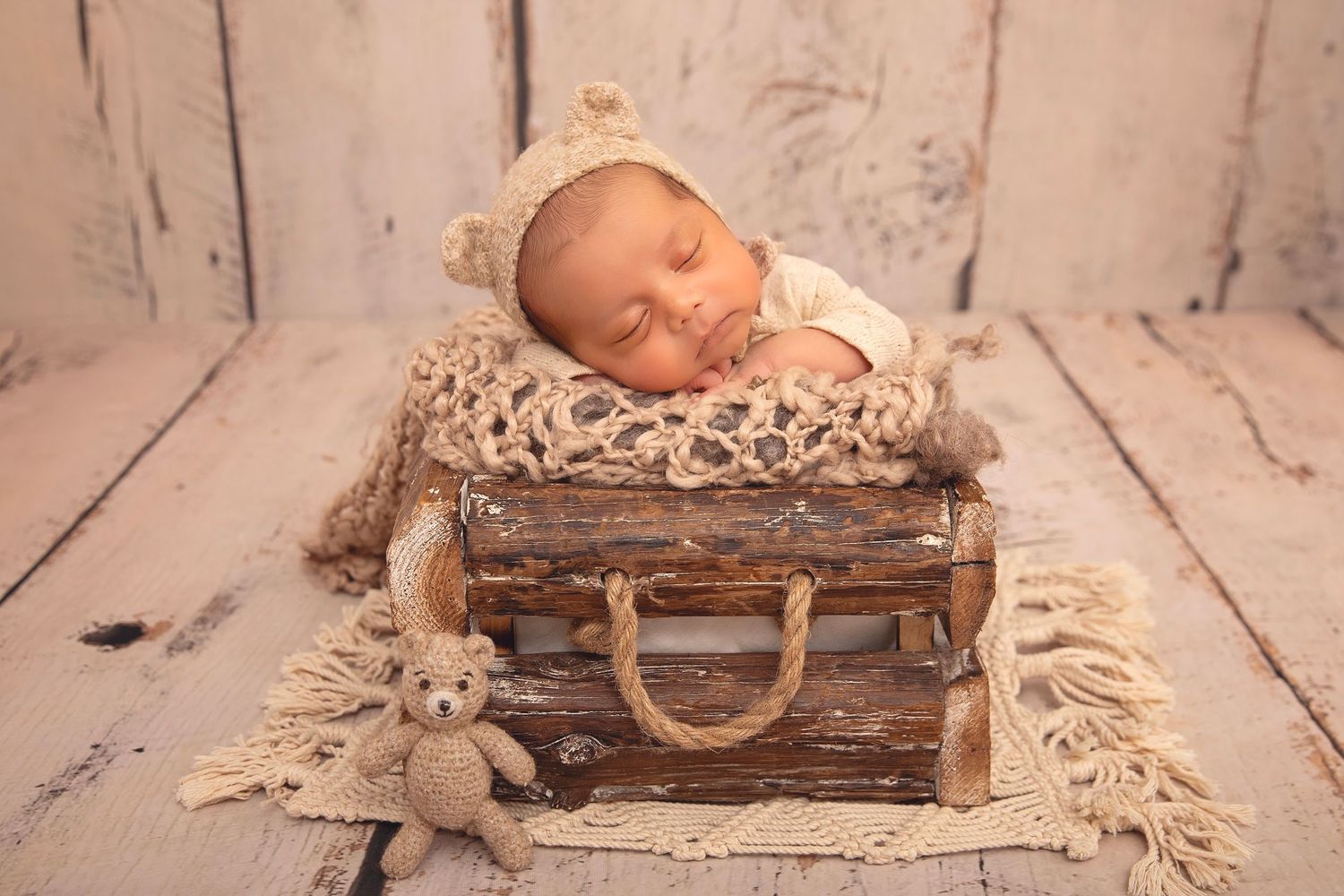 Sleeping newborn baby in bear hat nestled in rustic wooden suitcase with knit blanket and teddy bear.