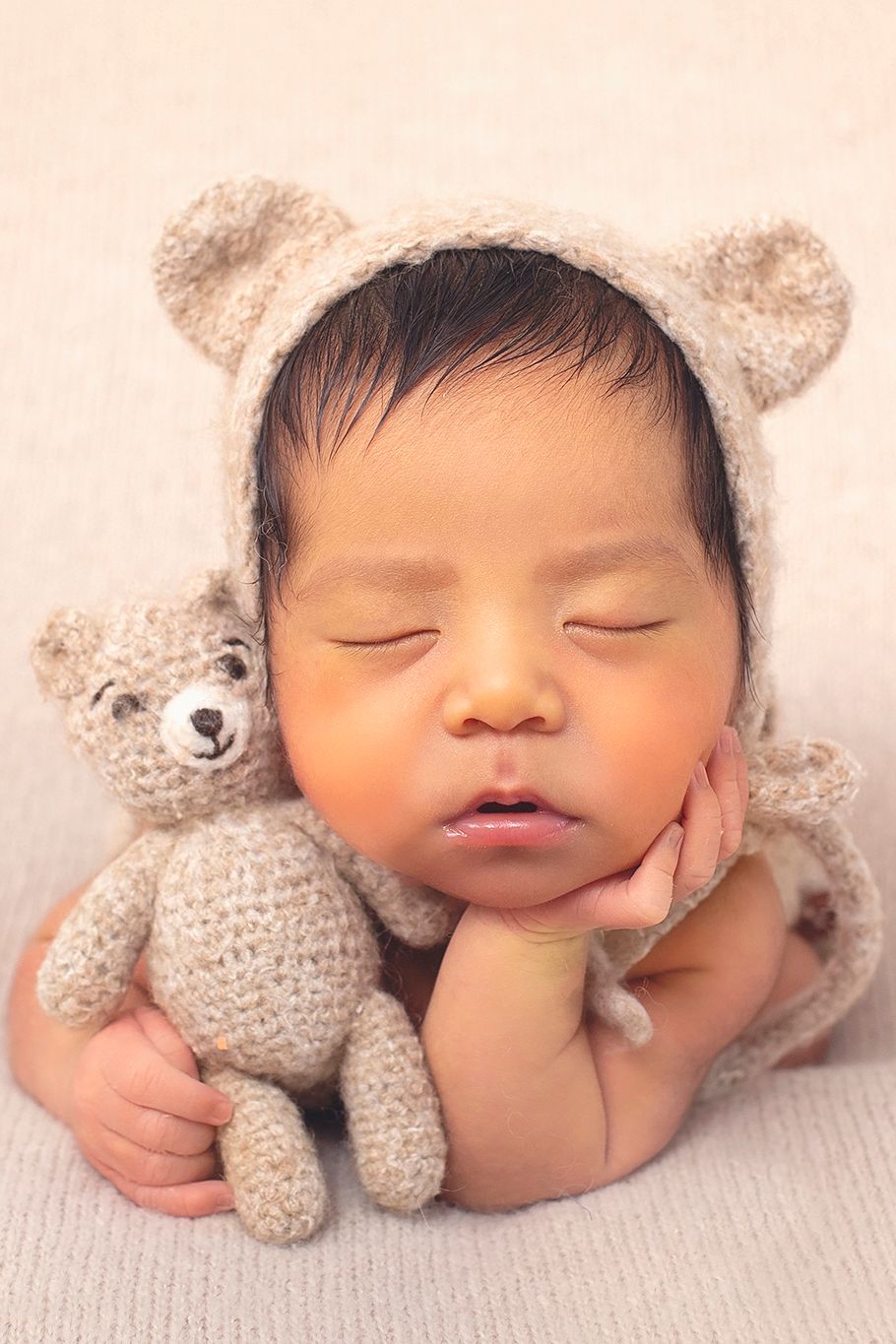 Sleeping newborn baby wearing a knit bear bonnet cuddles a small crocheted teddy bear on a soft beige blanket.