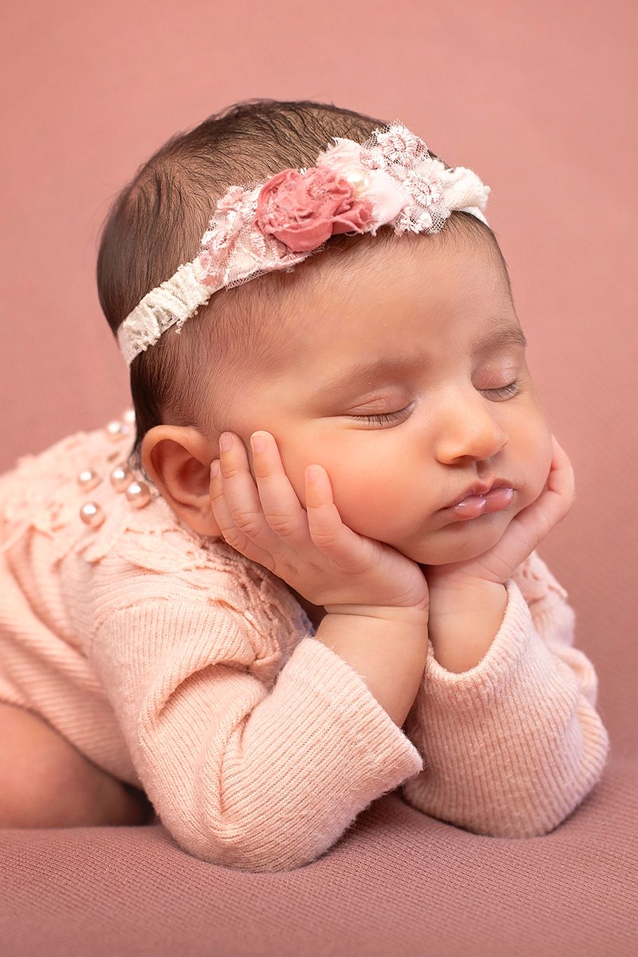 Sleeping newborn baby girl in pink outfit and floral headband, resting cheeks on hands in an adorable posed portrait.
