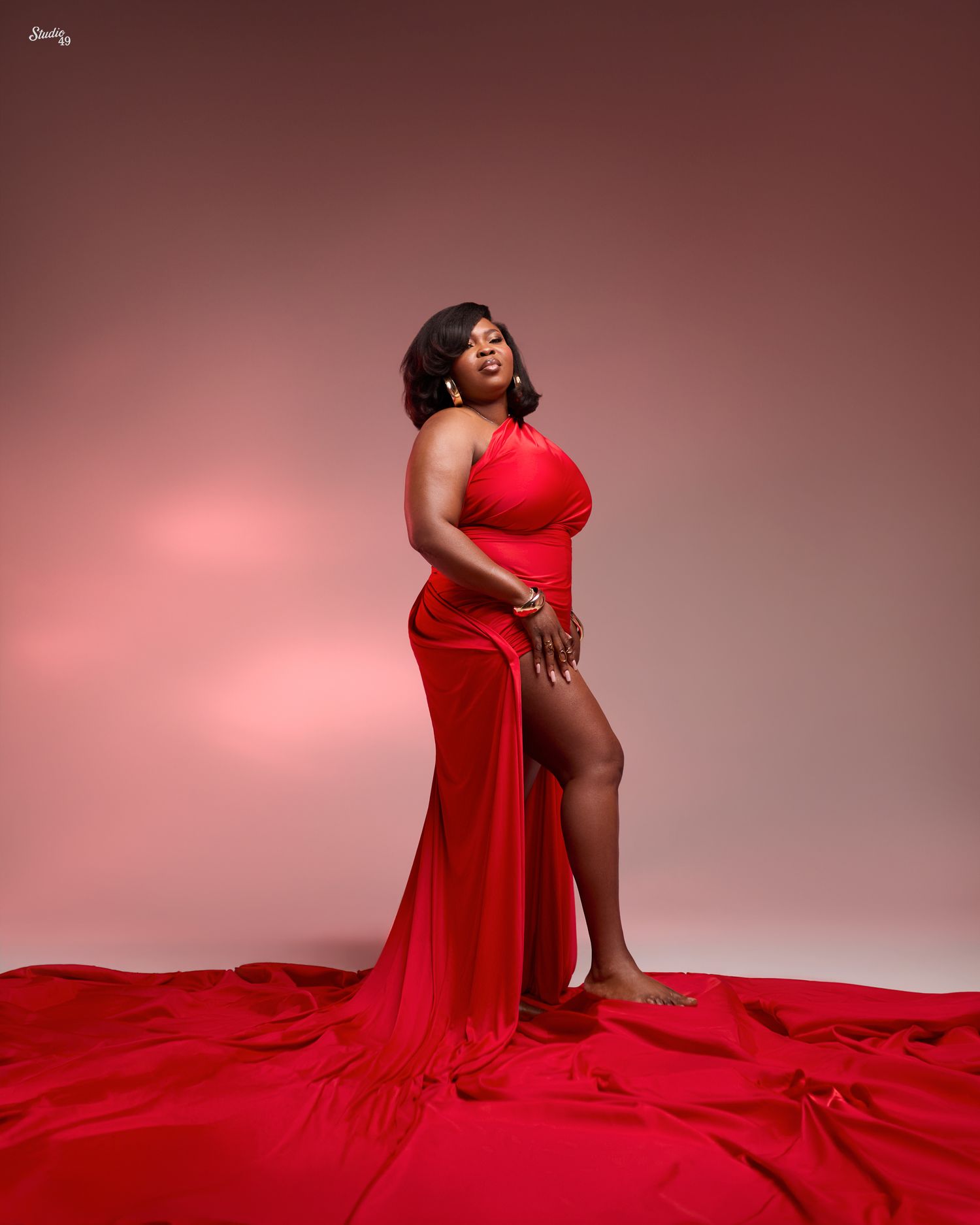 Woman poses confidently in a flowing red halter dress with a high slit, her train spread across the floor against a pink studio backdrop.