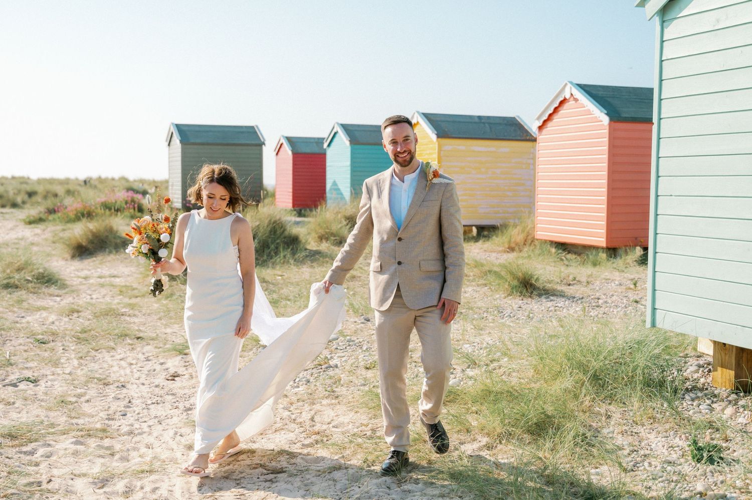 Wedding couple walking between colorful beach huts on sandy shore.