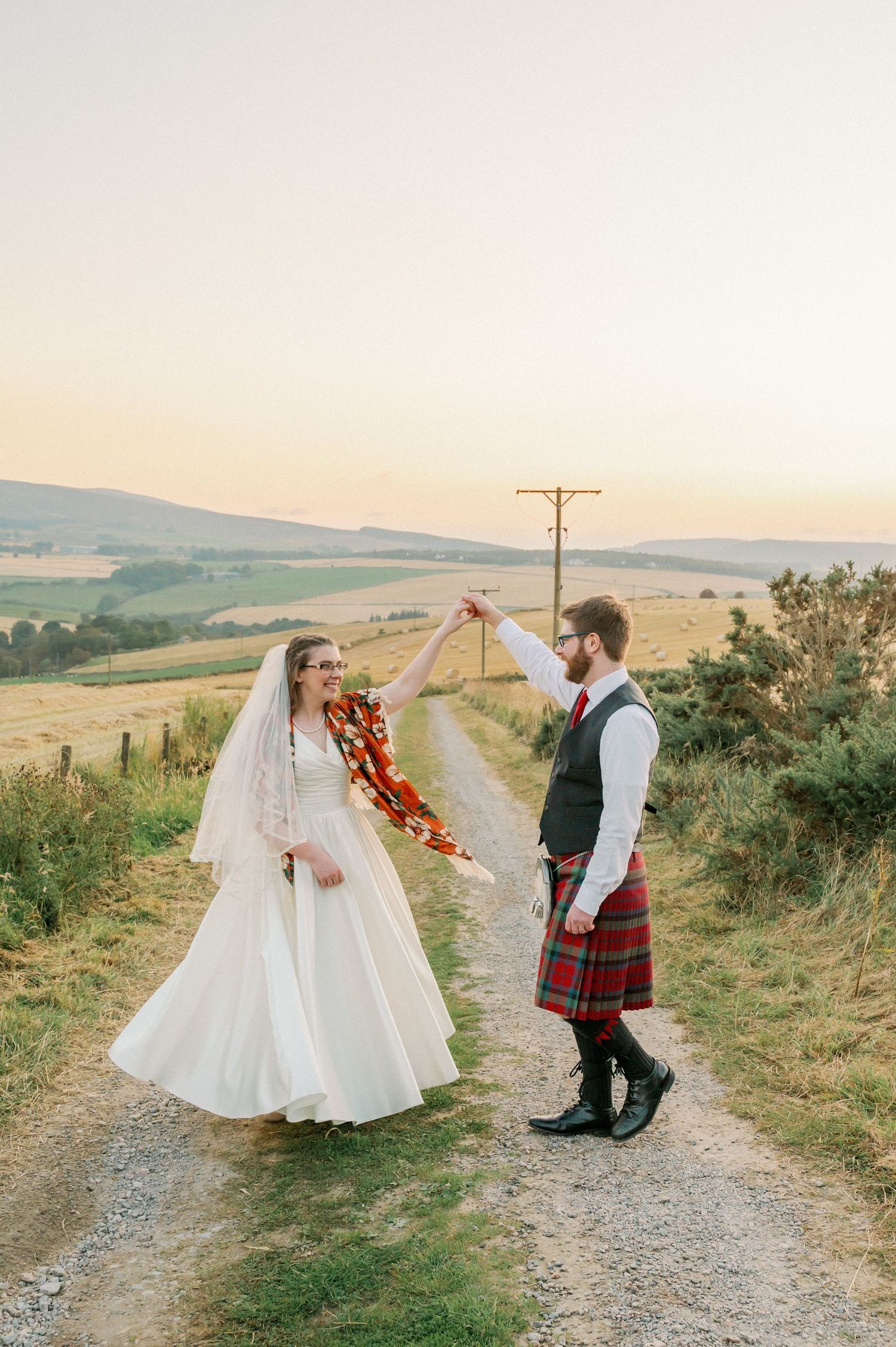 A romantic wedding couple walks along a scenic country path with the bride wearing white and groom in traditional Scottish kilt.