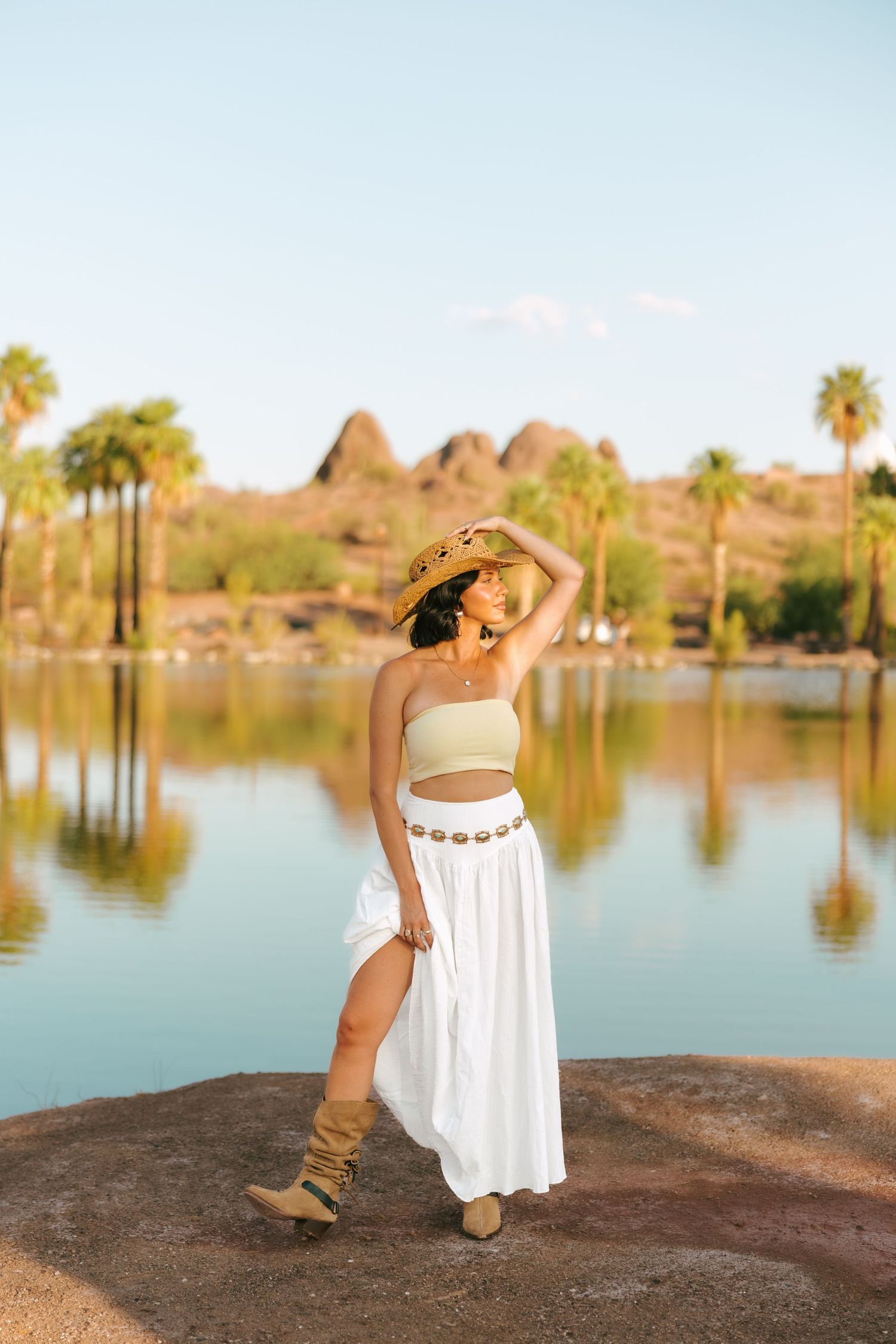 A person in a white dress poses gracefully by a serene lake surrounded by palm trees and mountains at sunset.
