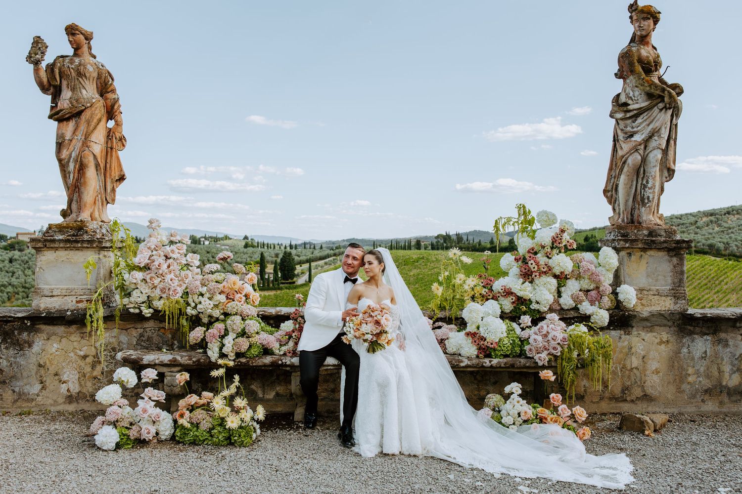 Elegant wedding dress displayed against stone wall with floral arrangements and garden statues in countryside setting.
