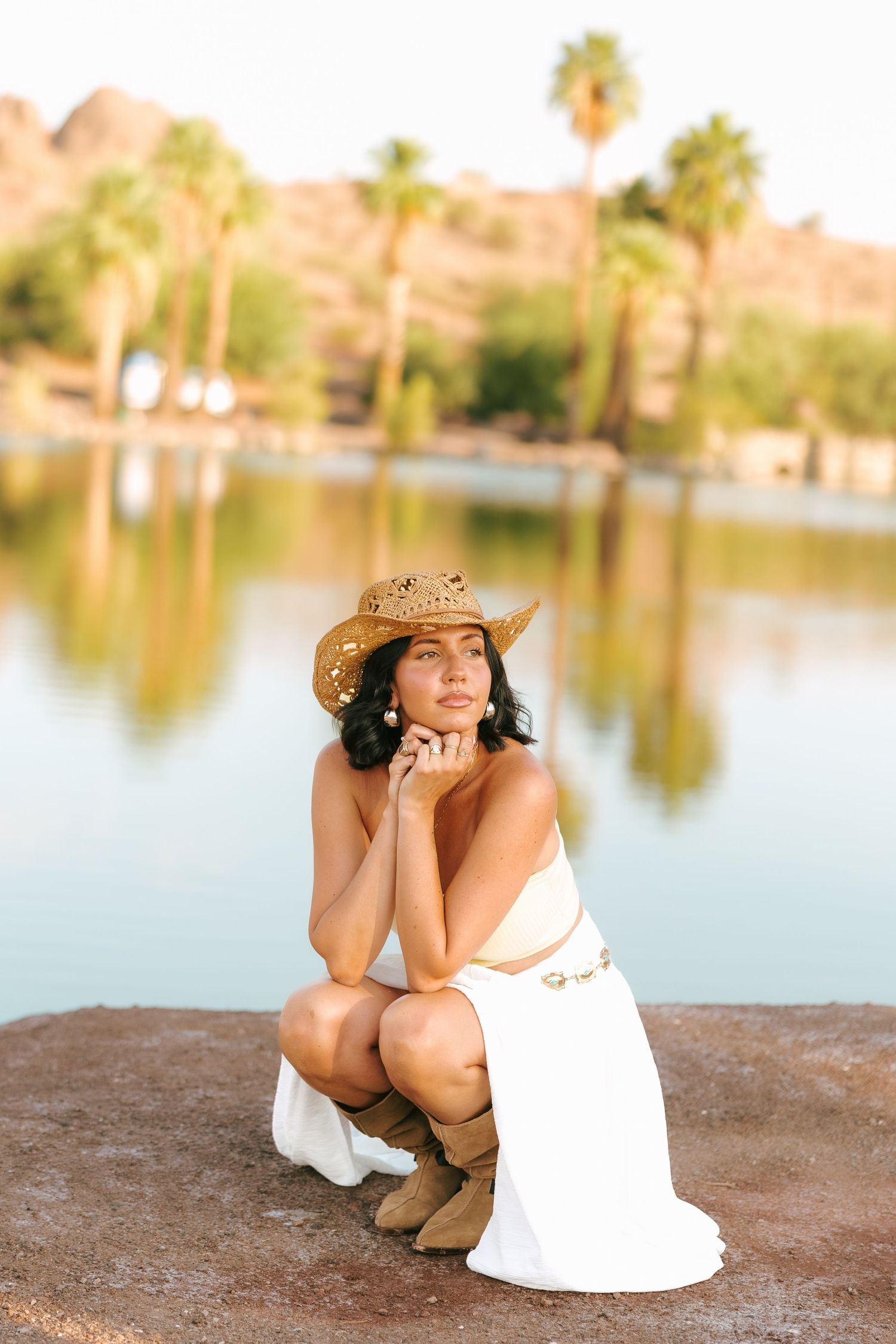 A person in a white dress and cowboy hat poses by a serene lake during golden hour.