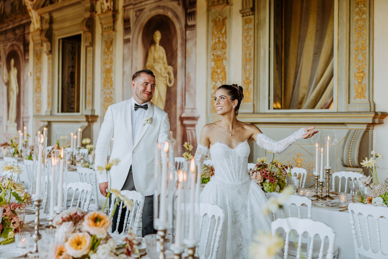 Elegant wedding reception table settings in an ornate white ballroom with crystal chandeliers and floral centerpieces.