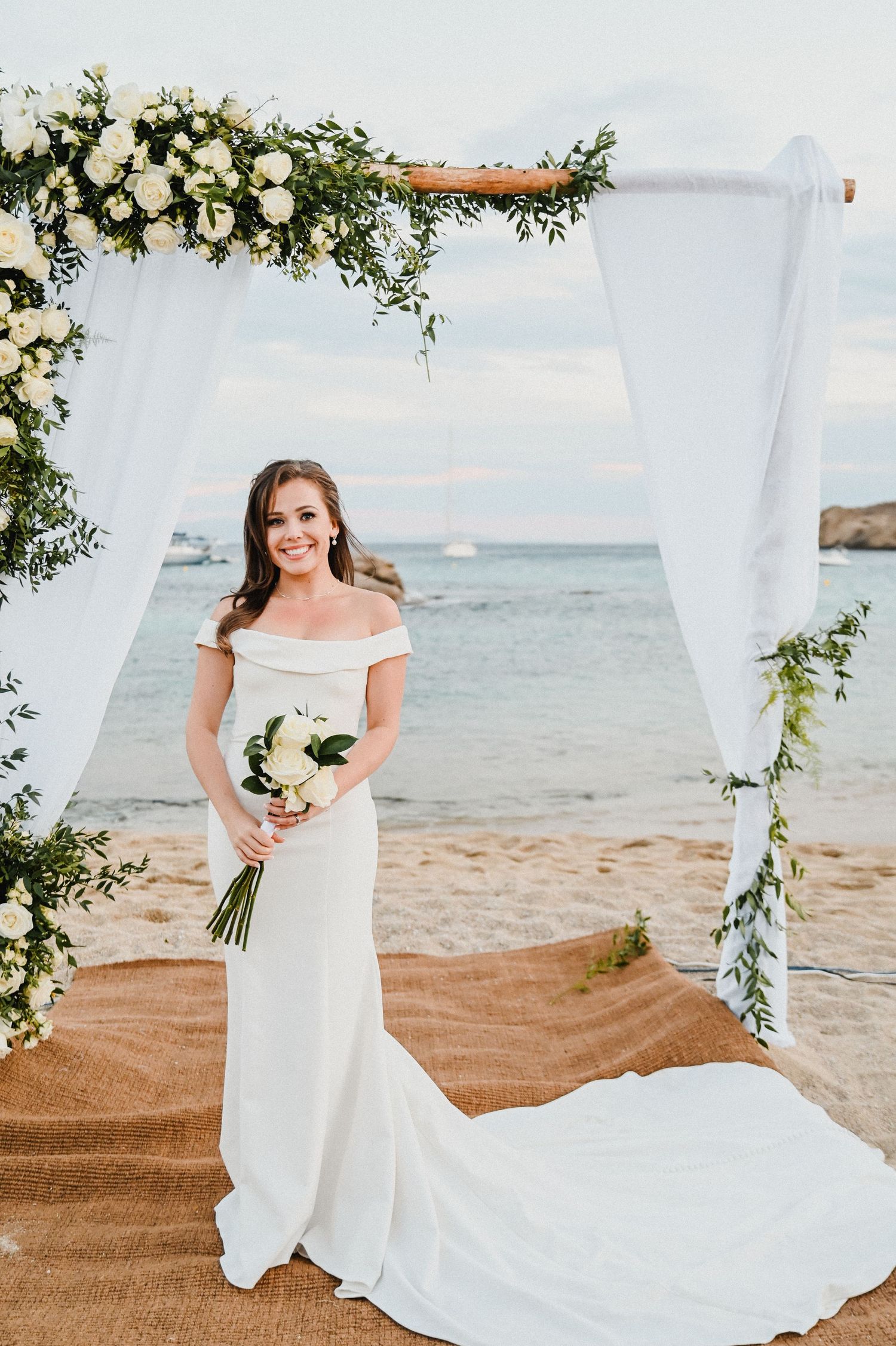 Beachside wedding with white draped arch adorned with roses and greenery overlooking the ocean.