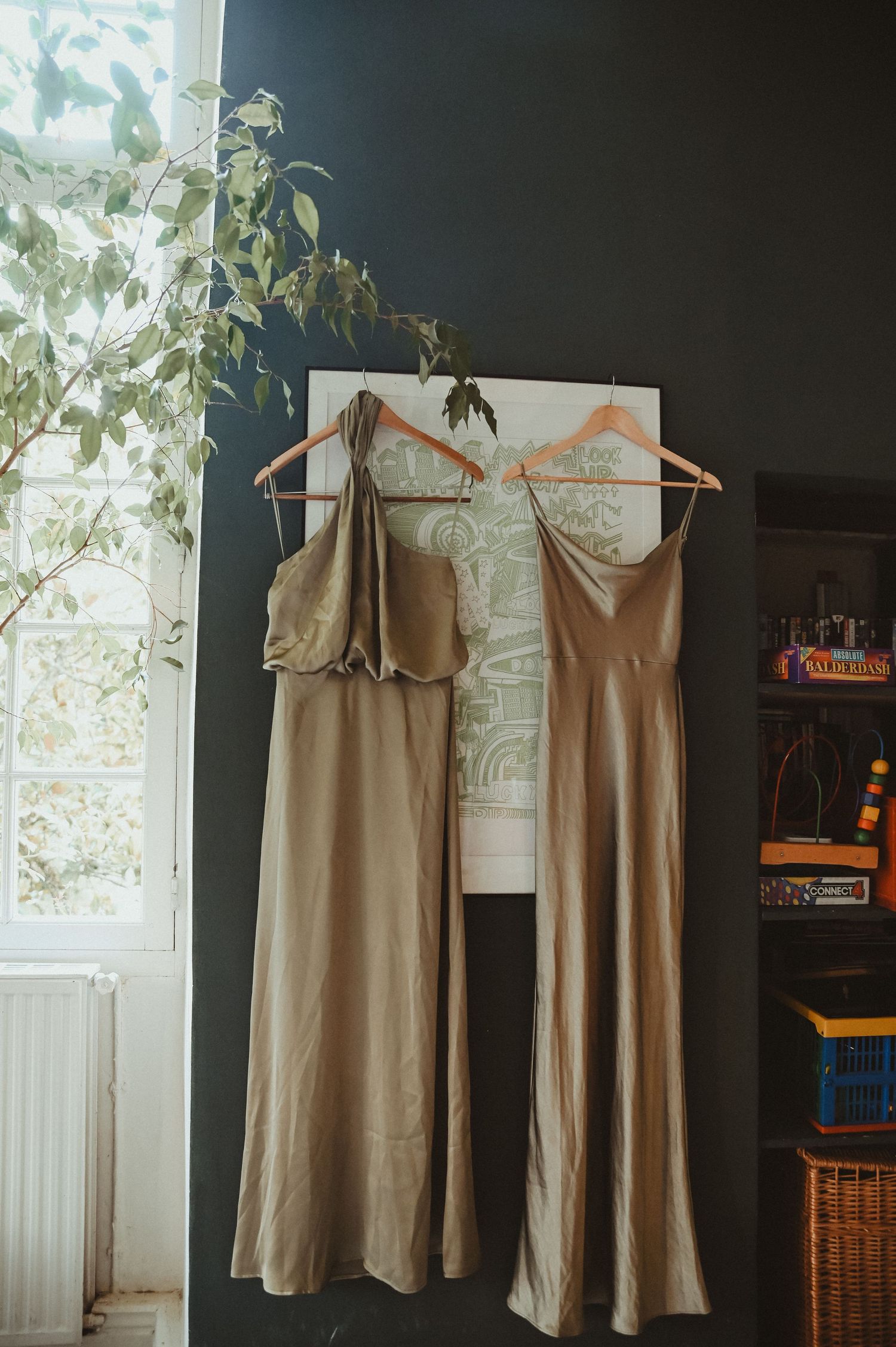 Two elegant silk bridesmaid dresses hanging by a window with natural light streaming in.