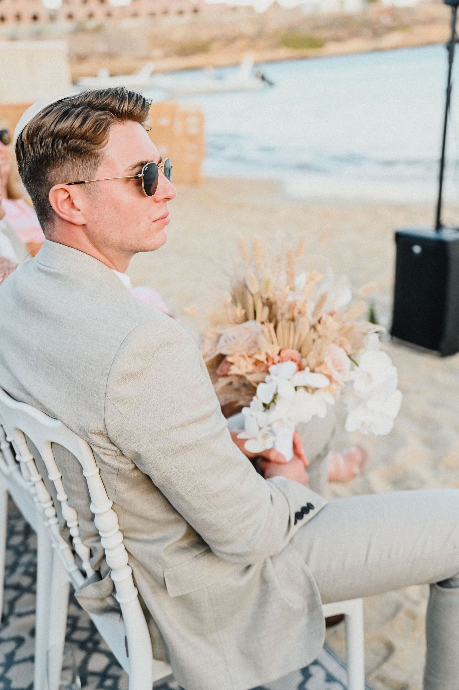 Wedding guest in beige suit and sunglasses seated in white chair by coastal backdrop.