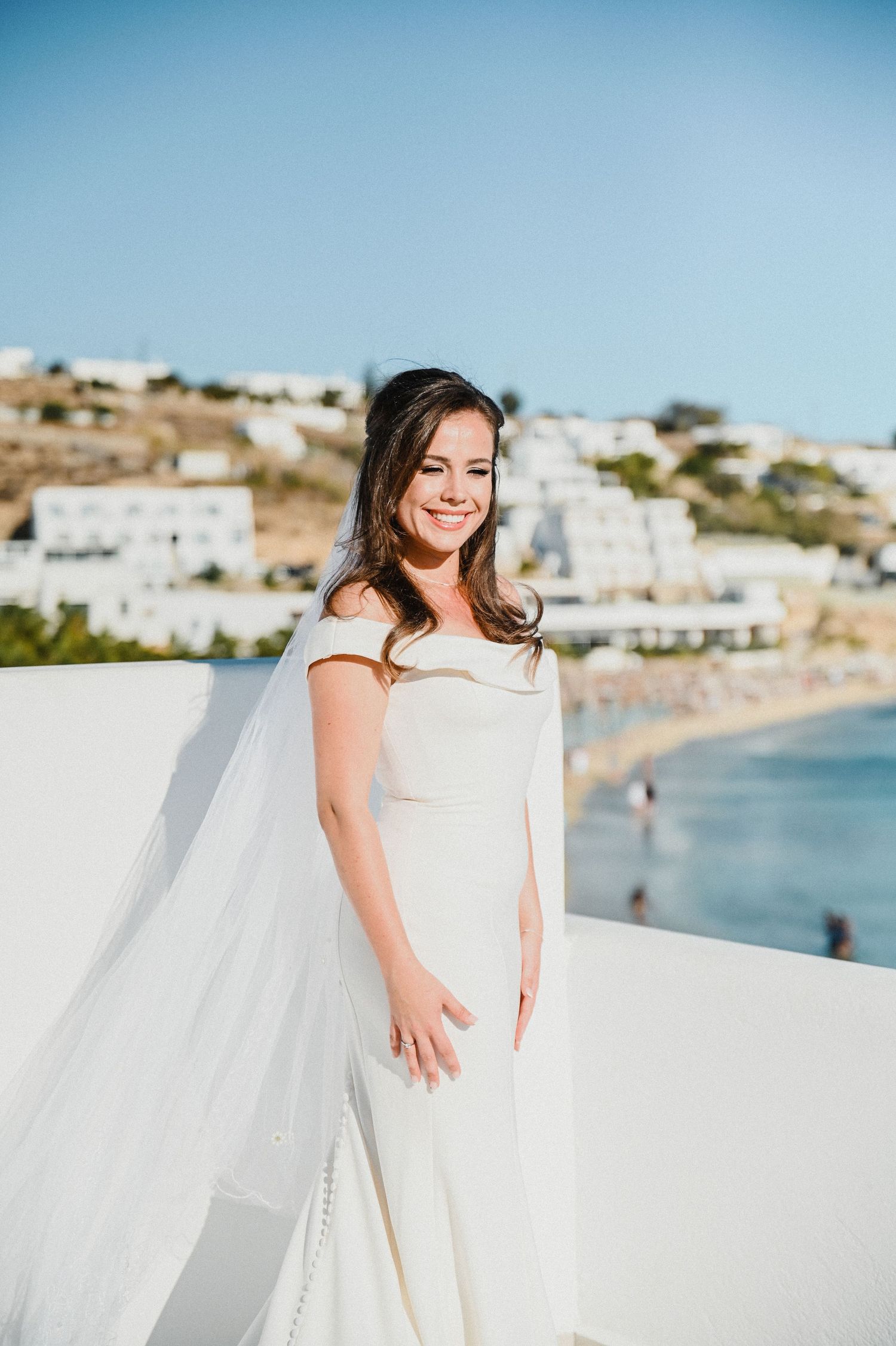 Bride in off-shoulder white dress smiles on terrace with coastal village backdrop.