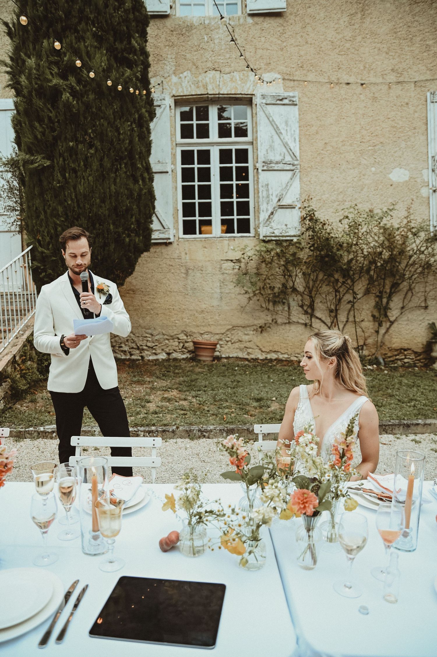 Wedding reception speech at elegant outdoor dinner with floral arrangements and white tablecloths.
