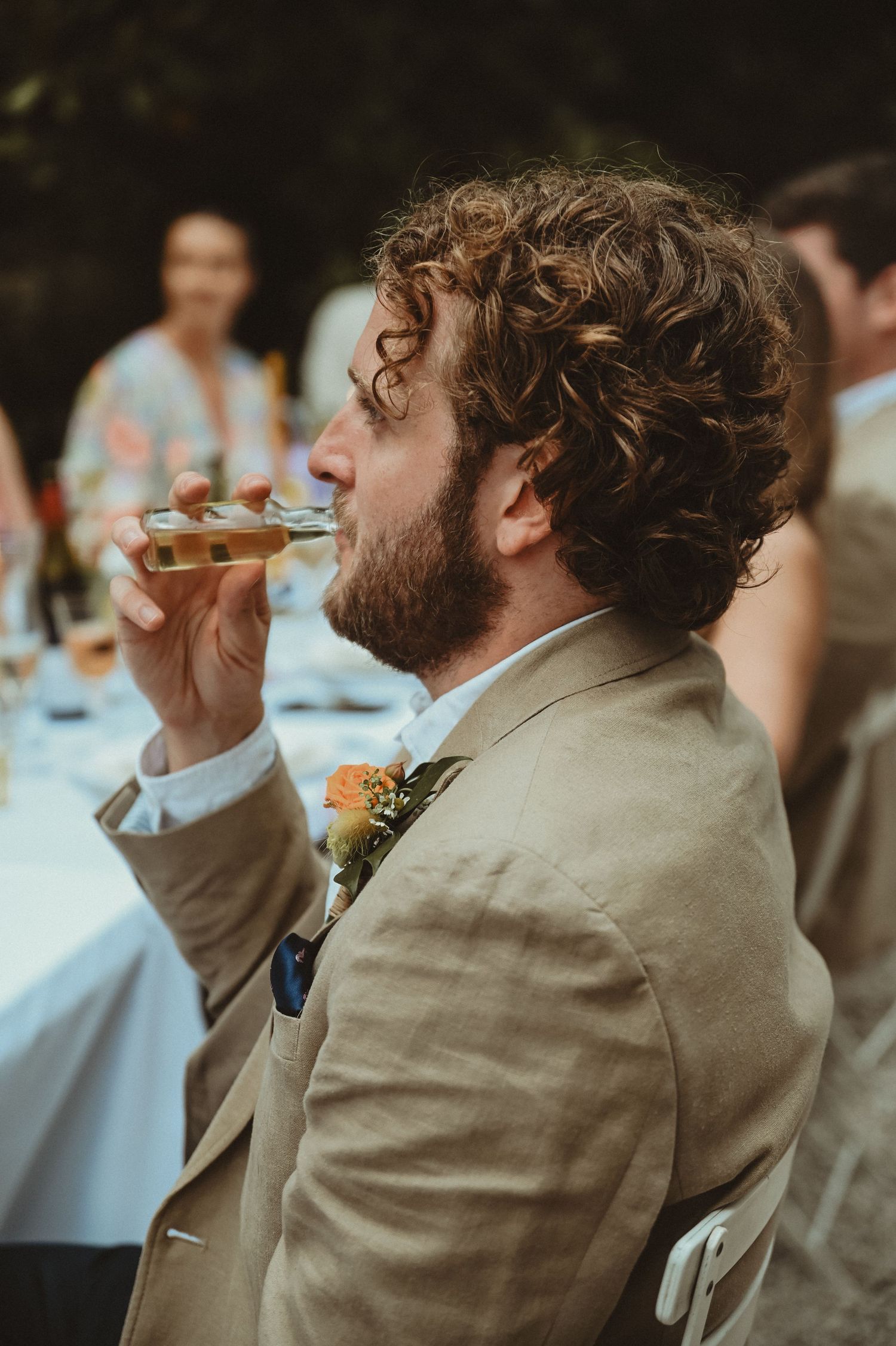 Guest in beige suit takes a sip from champagne glass during outdoor wedding reception.