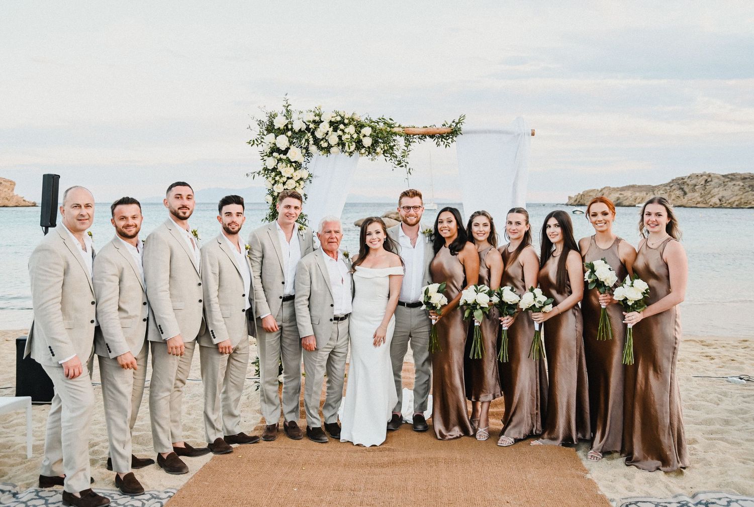 Wedding party posing under a floral arch on a beach with bridesmaids in brown dresses.