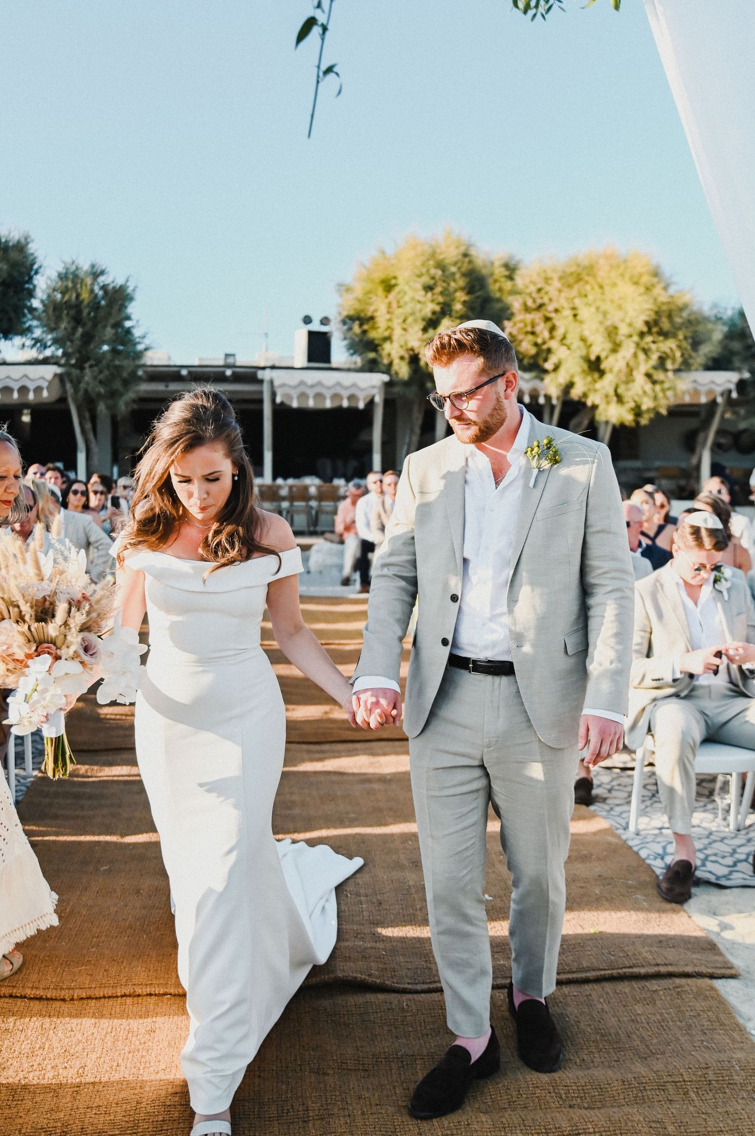 Newlyweds walking hand in hand on wooden deck during sunny outdoor ceremony.