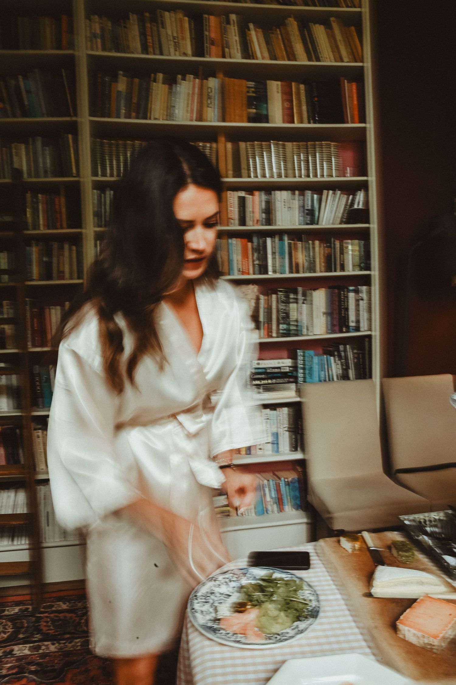Woman in white robe prepares food in a cozy home library setting with bookshelves in background.