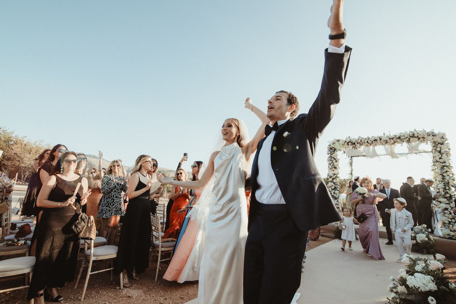 Newlyweds celebrate with raised arms during an outdoor wedding ceremony under a floral arch.