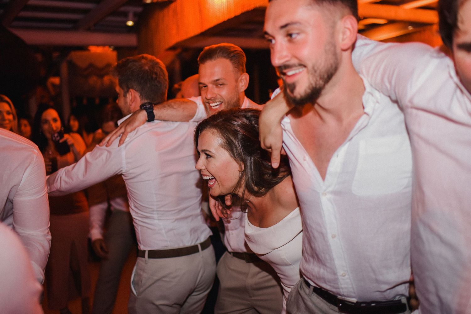 Wedding party members celebrate energetically on dance floor with orange ambient lighting.