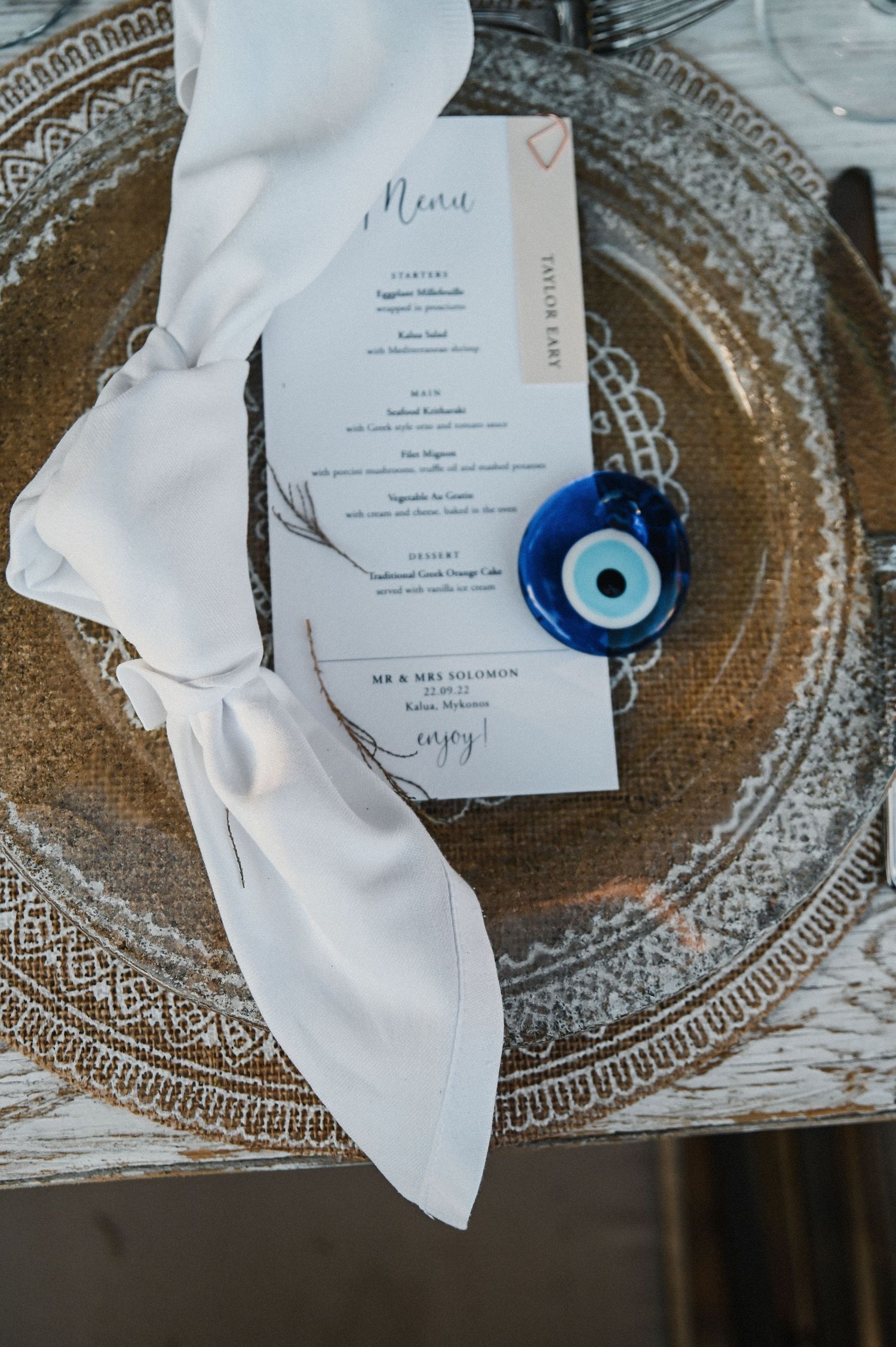 Wedding menu card with white ribbon and blue evil eye charm on ornate silver charger plate.