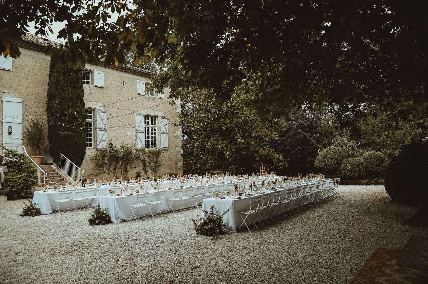 Elegant outdoor wedding venue with rows of white chairs set up on a gravel path beside a stone building.