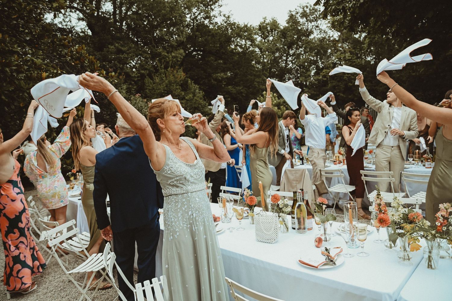 Wedding party celebrates with raised napkins around an outdoor table decorated with floral centerpieces.