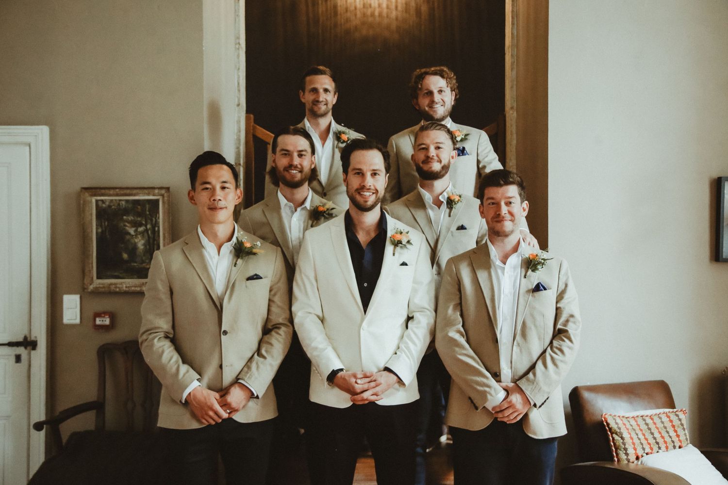 Groomsmen in beige suits pose with person in white jacket for indoor wedding portrait.