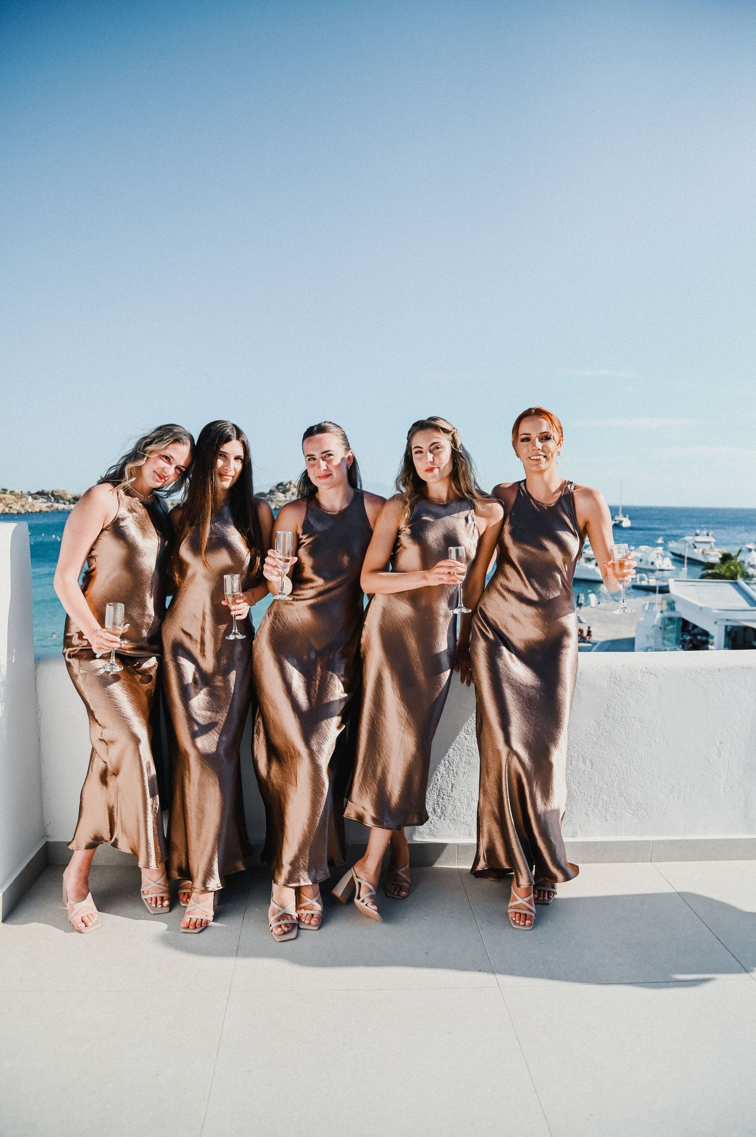 Bridesmaids in elegant champagne silk dresses pose together on a sunny rooftop terrace overlooking the ocean.