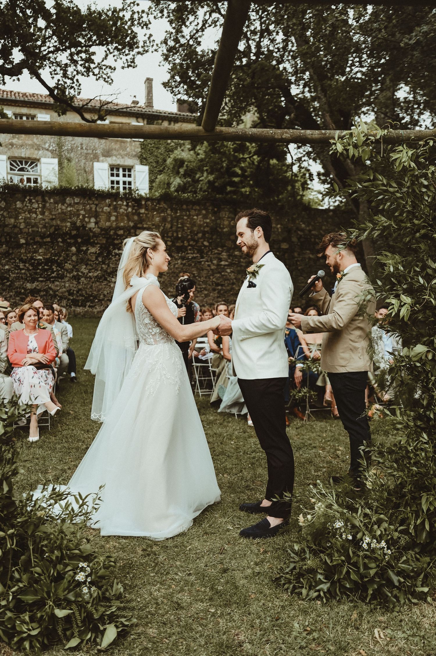 A wedding ceremony in a garden with stone walls and historic buildings in the background.