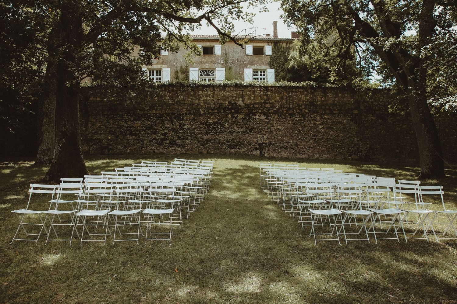Rows of white chairs set up for outdoor wedding ceremony with stone wall and building backdrop.