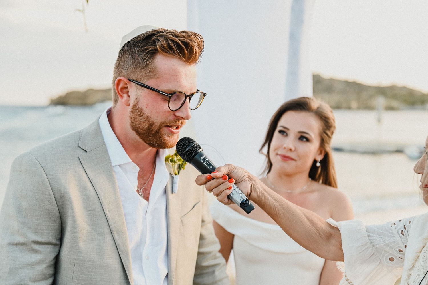 Couple shares a moment during ceremony speeches at a beachside wedding with microphone.