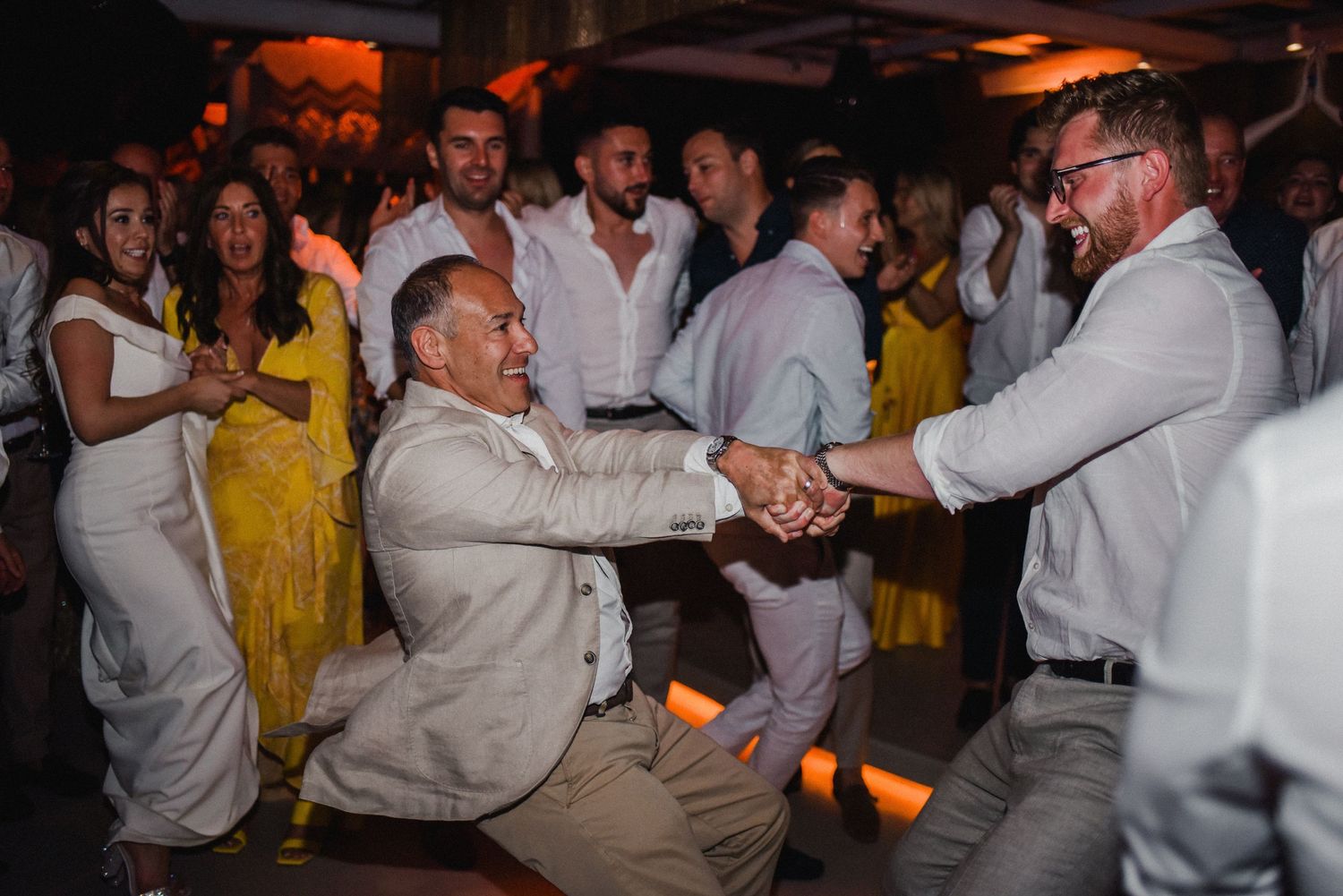 Wedding guests dancing together on reception dance floor with warm lighting.