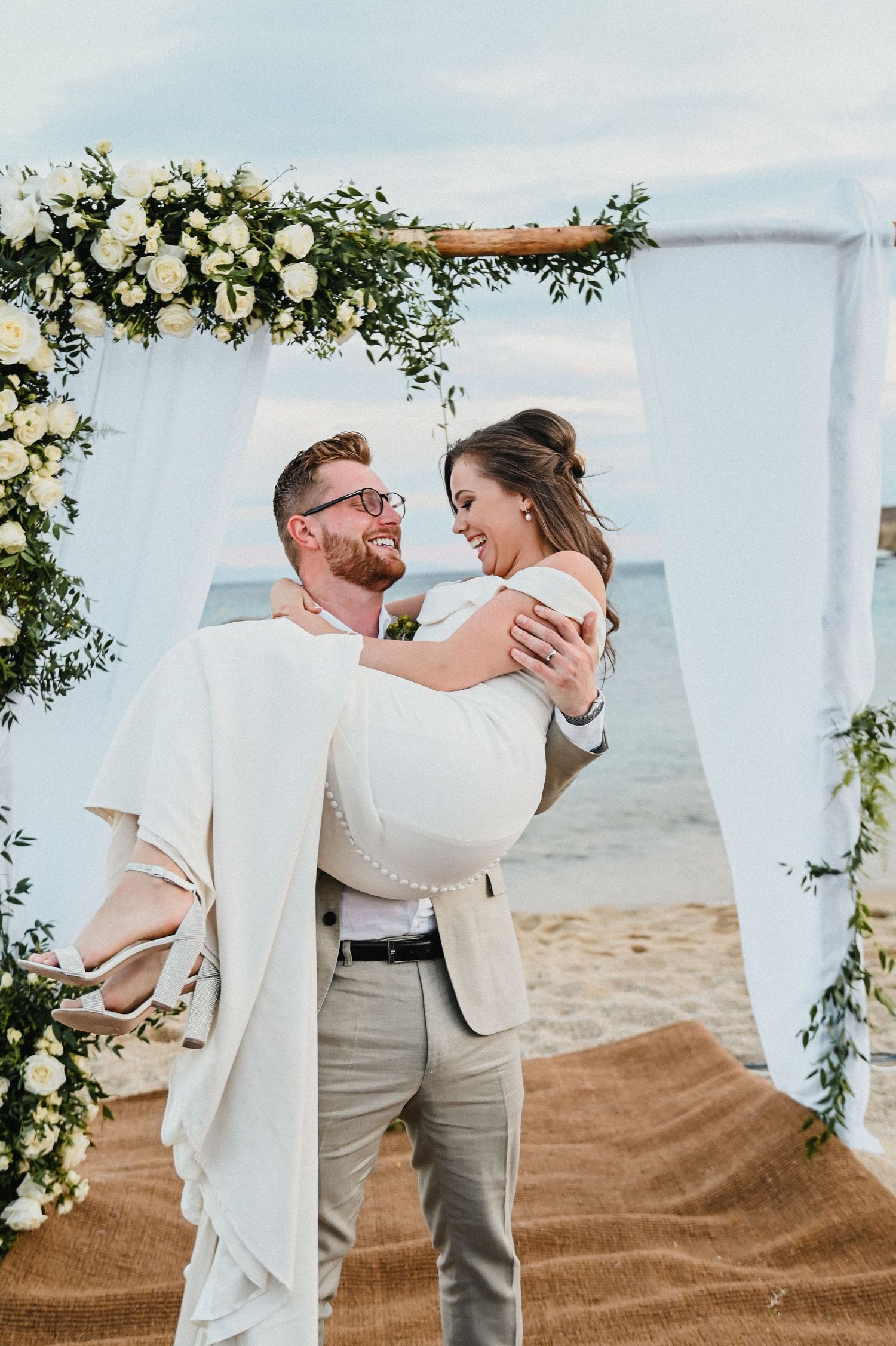A romantic beach wedding scene with a couple under a white fabric arch decorated with white flowers.