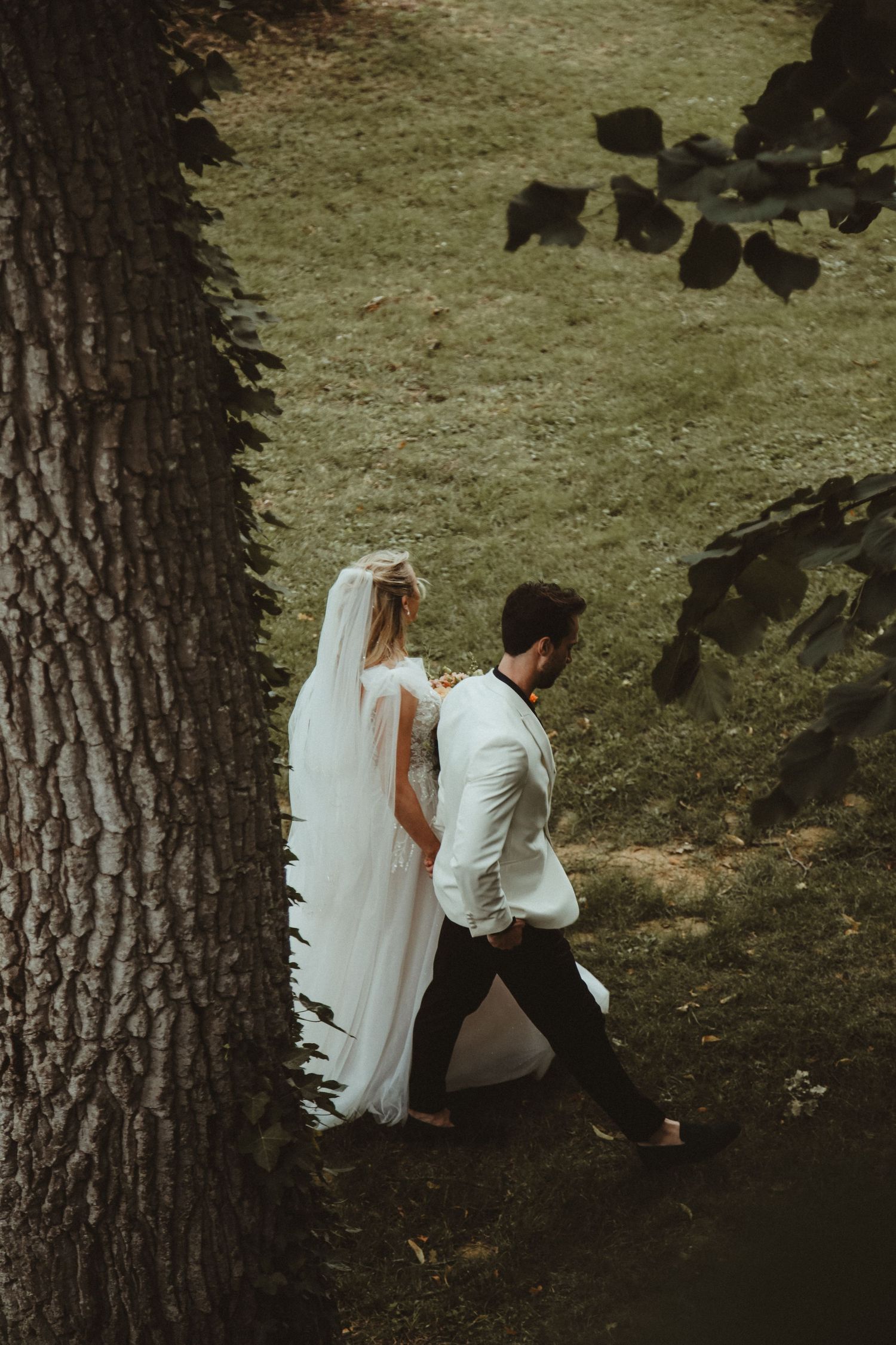 A wedding couple walks under the shade of a large tree in an outdoor garden setting.