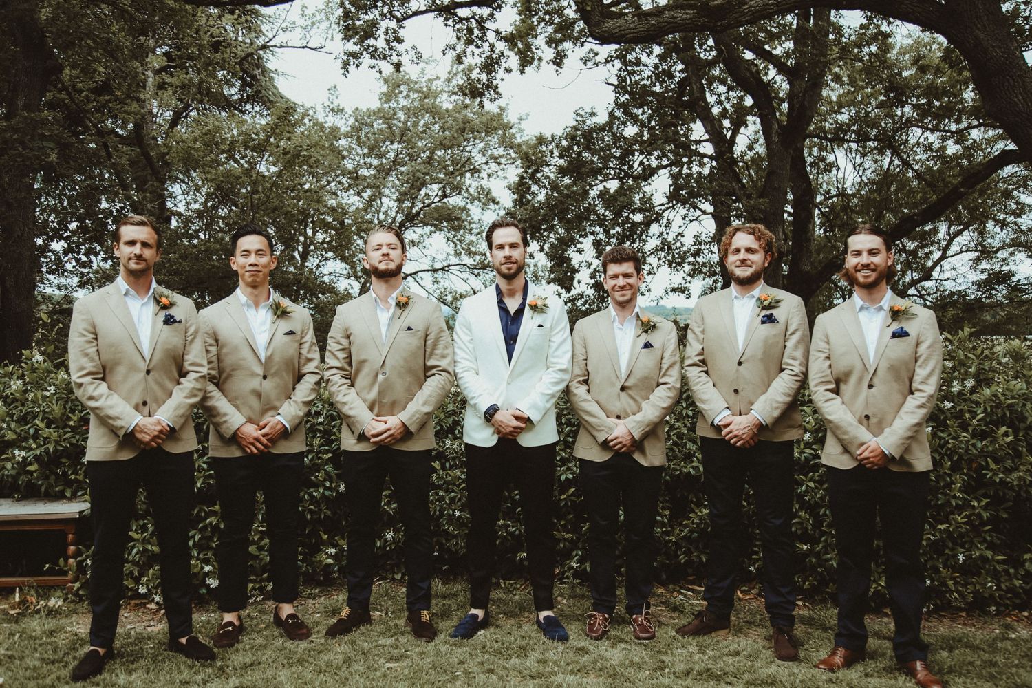 Wedding party in matching beige suits with one person in white stands outdoors under trees for formal group photo.