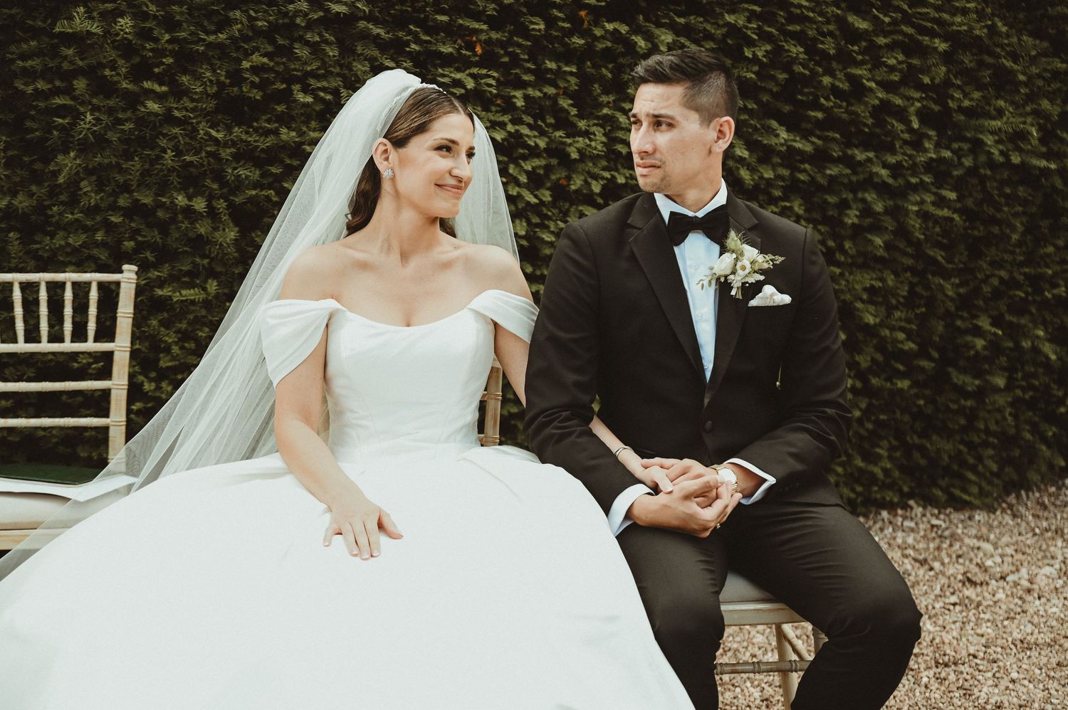 A wedding couple sits in an outdoor garden setting surrounded by classic architecture.
