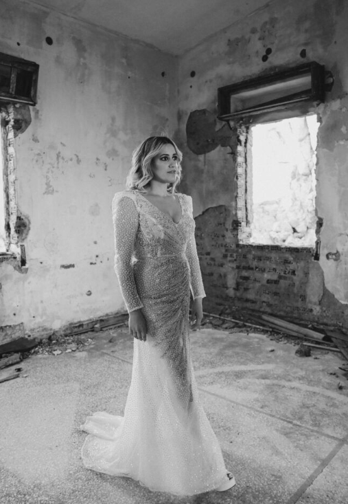 Elegant long-sleeved lace wedding dress photographed in a rustic room with distressed walls.