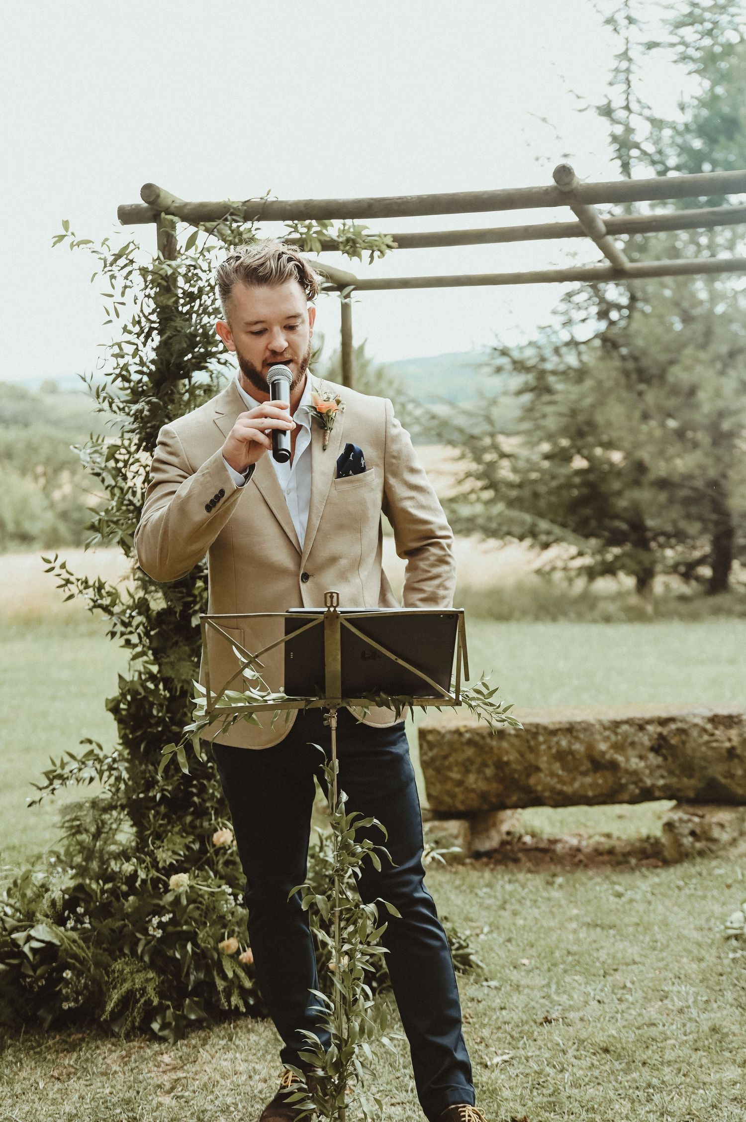 Groom in beige suit jacket reads from a box under wooden arch with climbing vines.