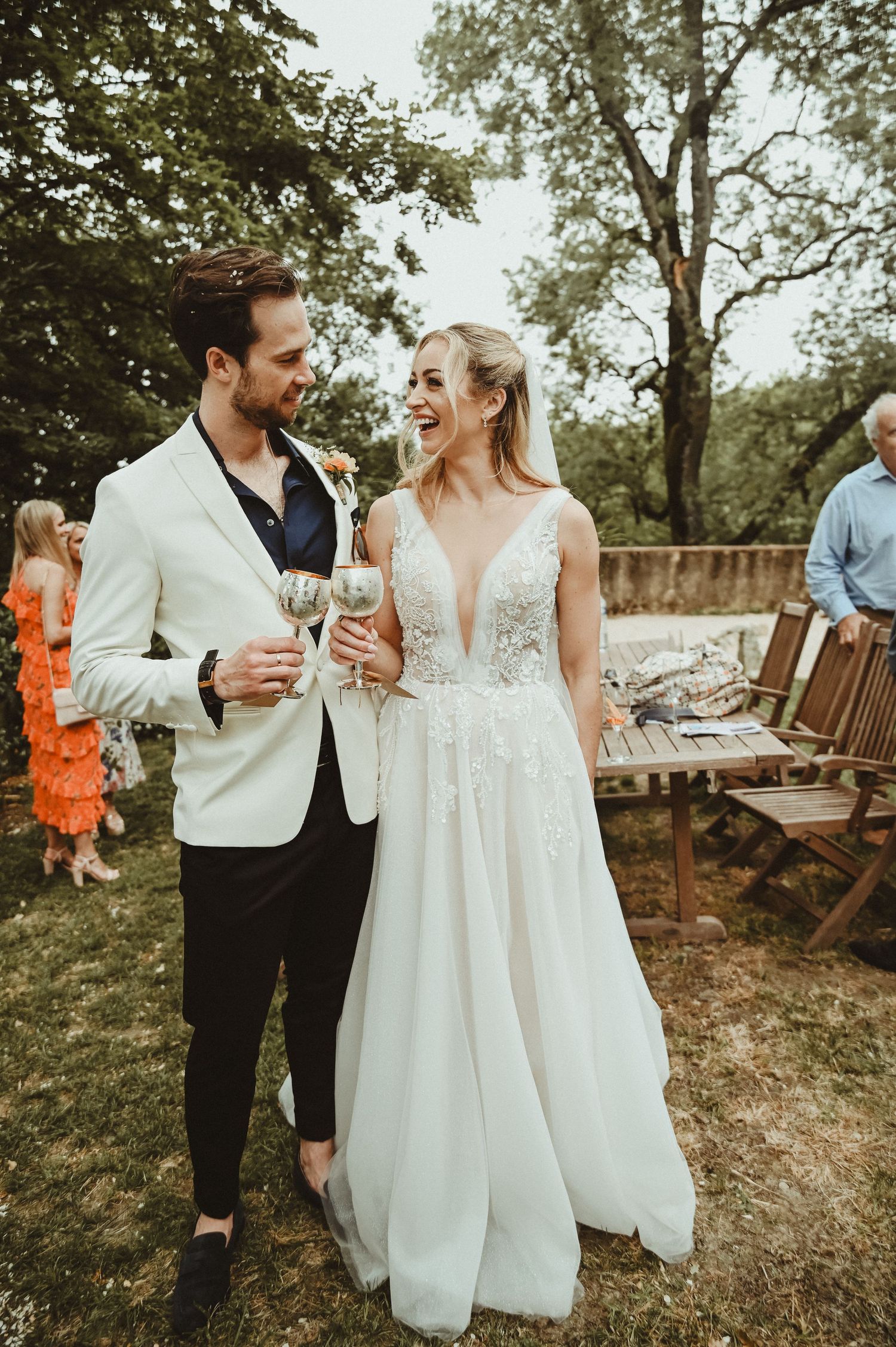 Couple in wedding attire share a laugh while holding champagne glasses at outdoor reception.