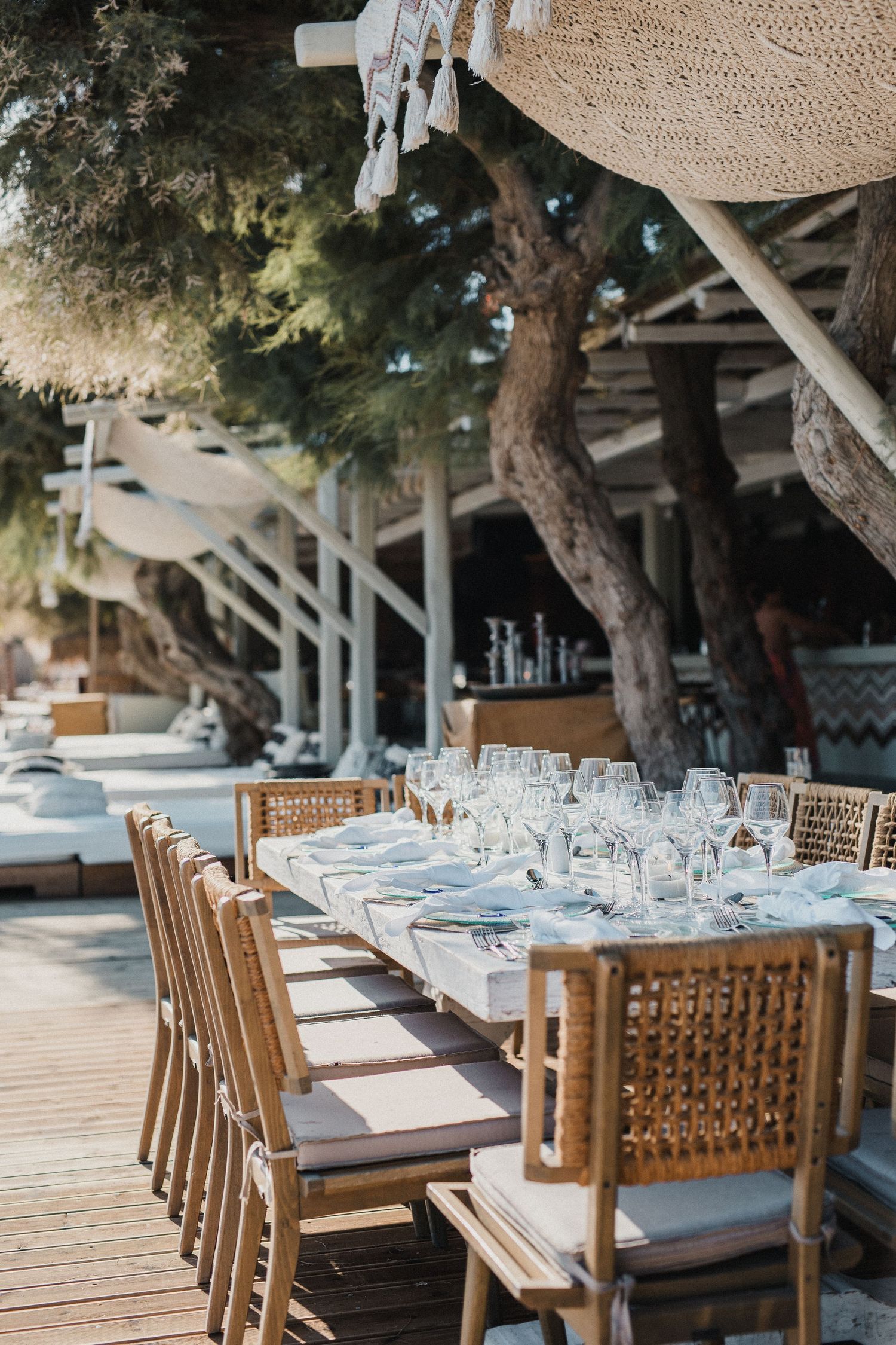 Rustic outdoor dining setup with wooden chairs and glassware under trees by the beach.