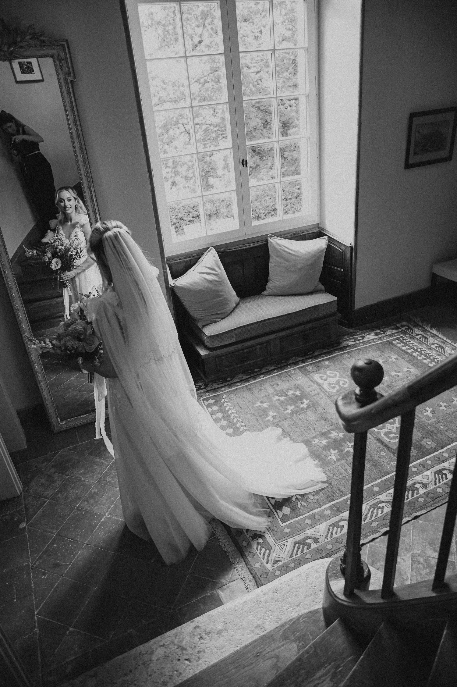 A flowing white wedding dress and veil cascades down stairs near a window and vintage loveseat.