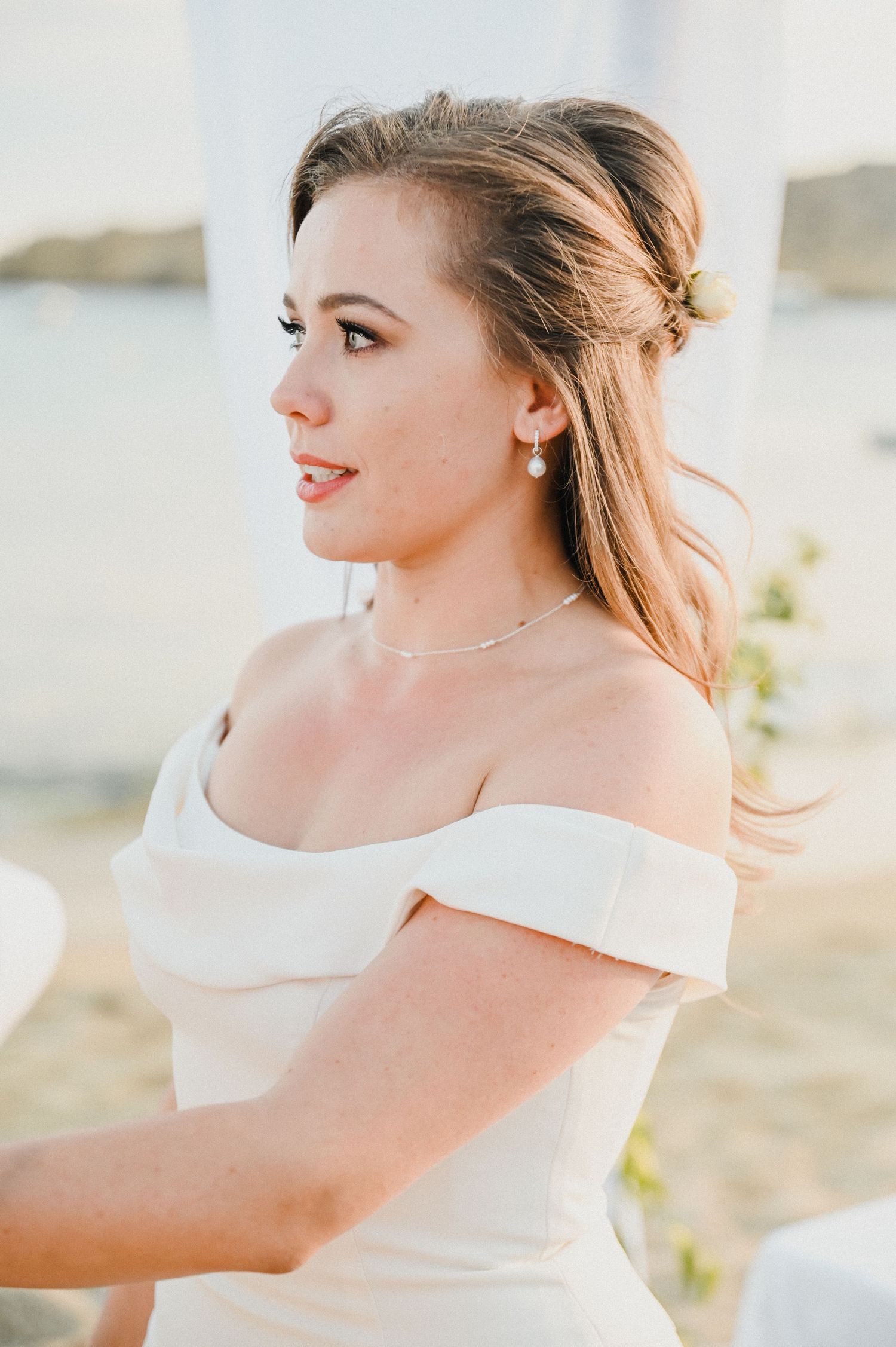 Bride in elegant off-shoulder gown with swept-back hair poses against coastal backdrop.