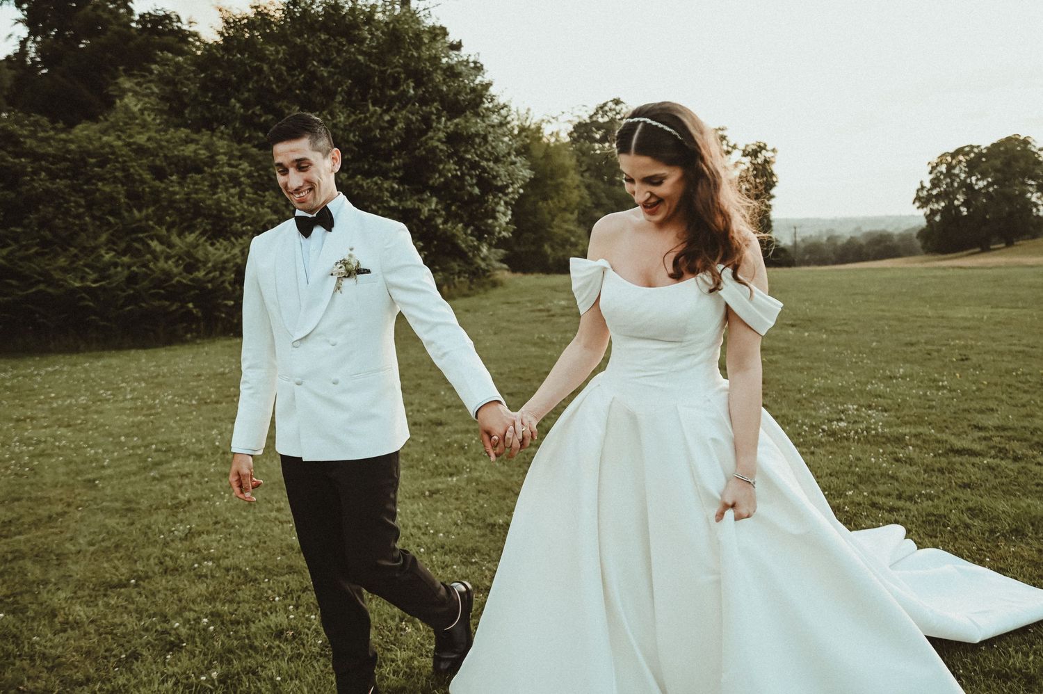 A couple walk hand in hand across a green lawn in elegant wedding attire.