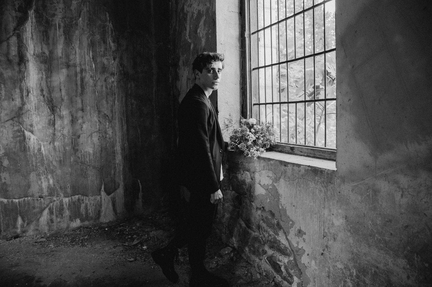 Black and white portrait of figure standing by industrial window holding floral arrangement.