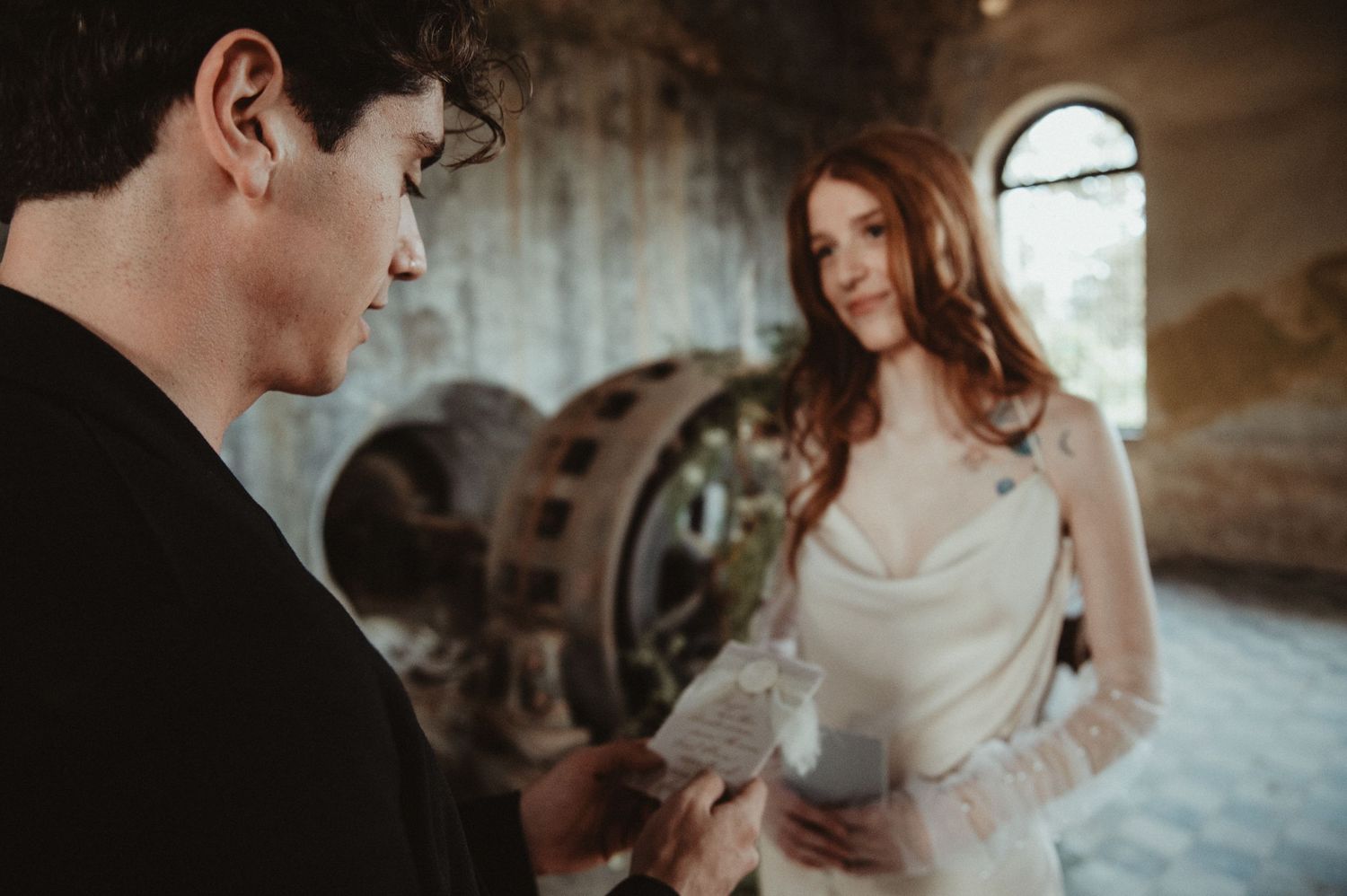An intimate wedding moment captured in a historic venue with arched windows and industrial elements.